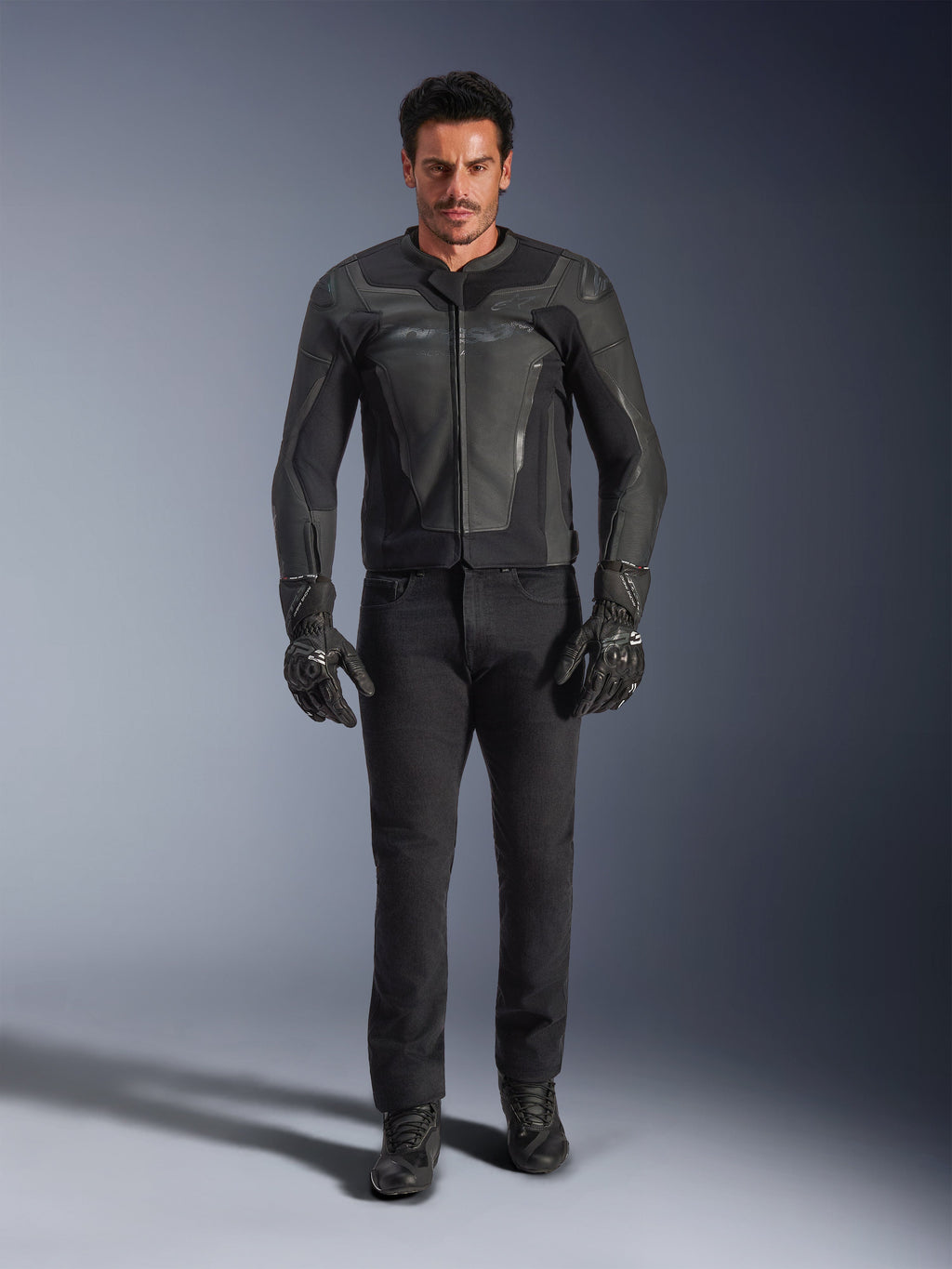 Chaqueta de cuero Alpinestars GP Force V2 en color negro, lucida por un piloto masculino con pantalones, guantes y botas de moto negros; construcción de cuero de estilo deportivo elegante con deslizadores en los hombros y protección integrada.