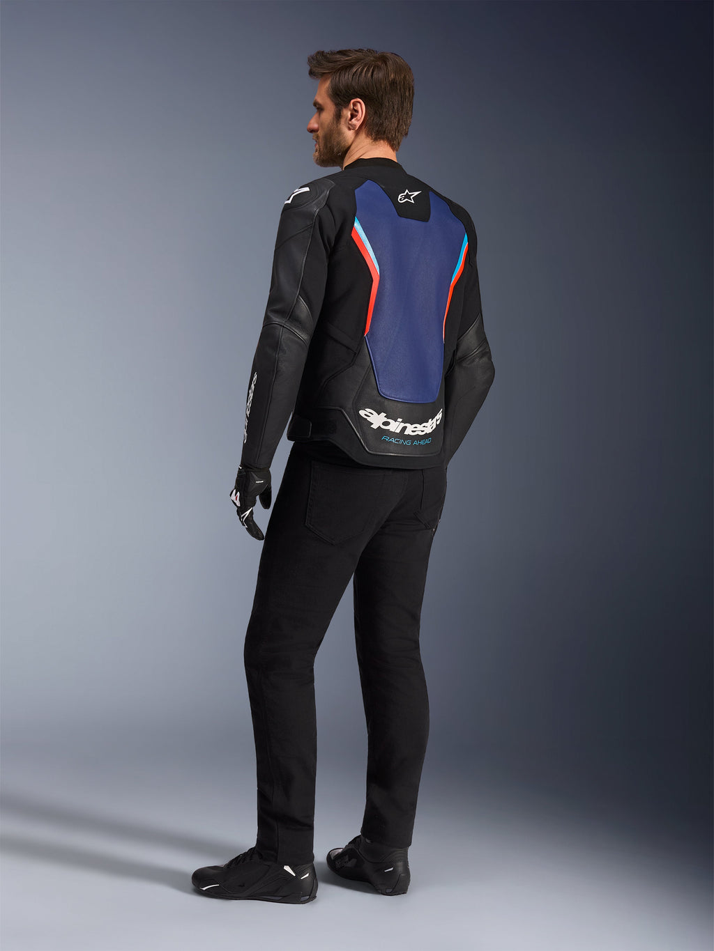 Vista trasera de tres cuartos de un modelo masculino vistiendo la chaqueta de cuero Alpinestars GP Force V2 en negro, azul y rojo flúor, resaltando el panel azul de la espalda con detalles en rojo fluorescente y logotipos blancos, combinada con pantalones negros y guantes de moto.
