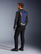 Vista trasera de tres cuartos de un modelo masculino vistiendo la chaqueta de cuero Alpinestars GP Force V2 en negro, azul y rojo flúor, resaltando el panel azul de la espalda con detalles en rojo fluorescente y logotipos blancos, combinada con pantalones negros y guantes de moto.
