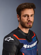 Chaqueta de cuero Alpinestars GP Force V2, negro, azul y rojo flúor, retrato en primer plano de un piloto masculino, con el logotipo blanco de Alpinestars en las protecciones de hombro DFS negras, panel de pecho de cuero azul con marca blanca y texto 'Racing Ahead', acentos en rojo flúor, equipamiento deportivo premium para motocicletas.