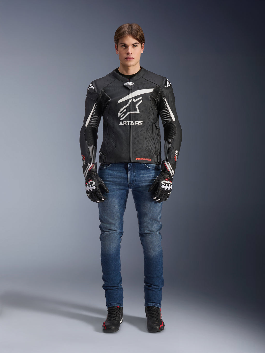 Modelo masculino vistiendo la chaqueta de cuero Alpinestars GP Plus R V4 Rideknit en negro y blanco, combinada con vaqueros de moto azules, guantes de moto negros y calzado de moto negro, con un gran logotipo blanco de Astars en el pecho, paneles de cuero perforado y franjas de carreras blancas en las mangas, vista frontal de cuerpo completo