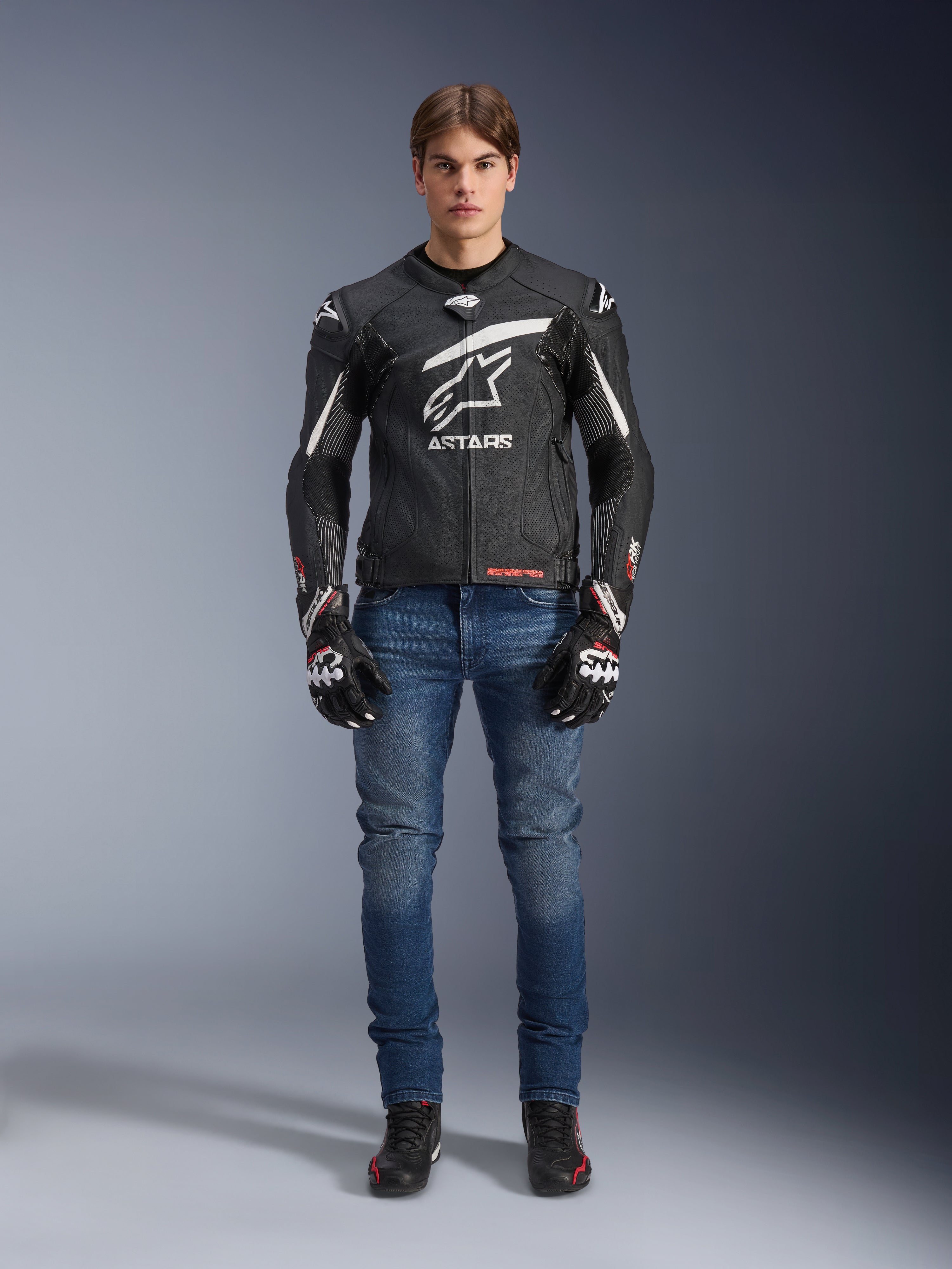 Modelo masculino vistiendo la chaqueta de cuero Alpinestars GP Plus R V4 Rideknit en negro y blanco, combinada con vaqueros de moto azules, guantes de moto negros y calzado de moto negro, con un gran logotipo blanco de Astars en el pecho, paneles de cuero perforado y franjas de carreras blancas en las mangas, vista frontal de cuerpo completo