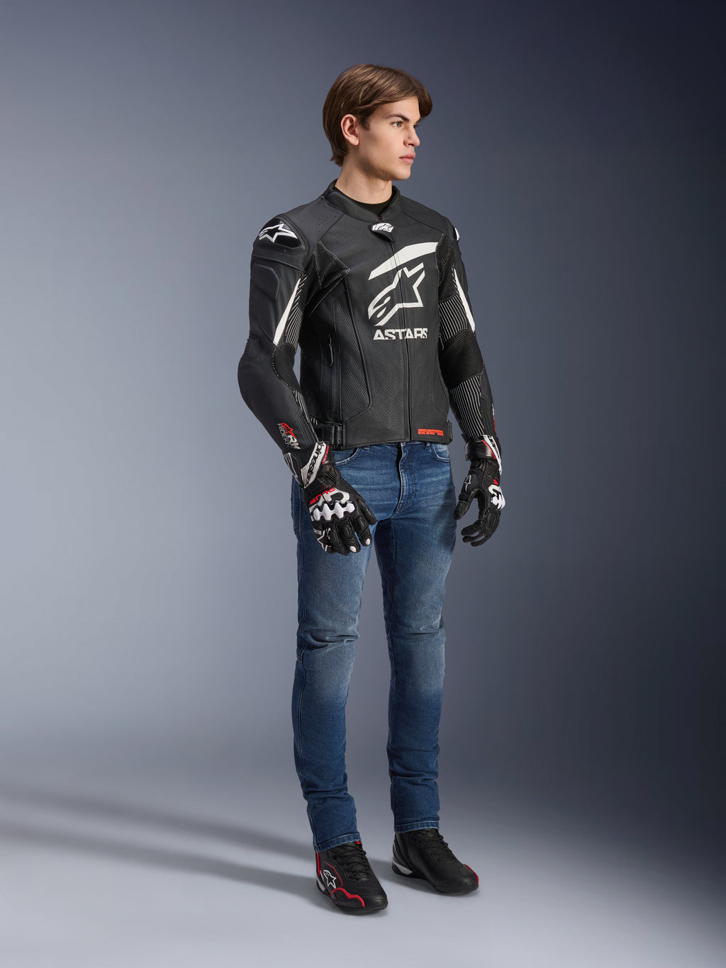 Alpinestars GP Plus R V4 Rideknit Leather Jacket, negro y blanco, chaqueta de cuero para moto vestida por un modelo con vaqueros azules y guantes negros, con un prominente logotipo blanco de Astars en el pecho, paneles perforados y detalles blancos en los hombros.