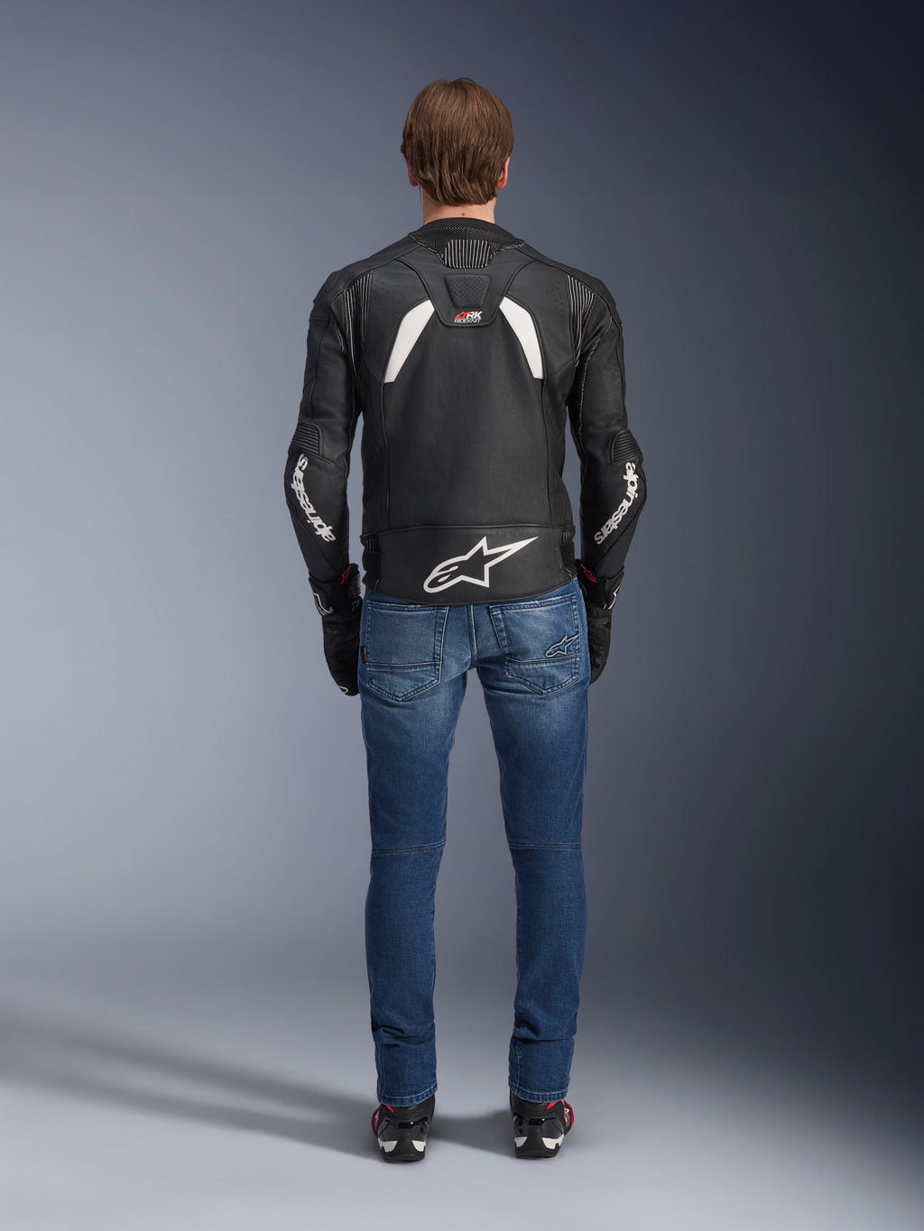 Vista trasera de la chaqueta de cuero Alpinestars GP Plus R V4 Rideknit, en color negro y blanco, con un logotipo blanco grande en la parte baja de la espalda y marca en las mangas, usada con vaqueros azules y botas negras, destacando su construcción técnica y ajuste deportivo.