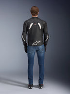 Vista trasera de la chaqueta de cuero Alpinestars GP Plus R V4 Rideknit, en color negro y blanco, con un logotipo blanco grande en la parte baja de la espalda y marca en las mangas, usada con vaqueros azules y botas negras, destacando su construcción técnica y ajuste deportivo.