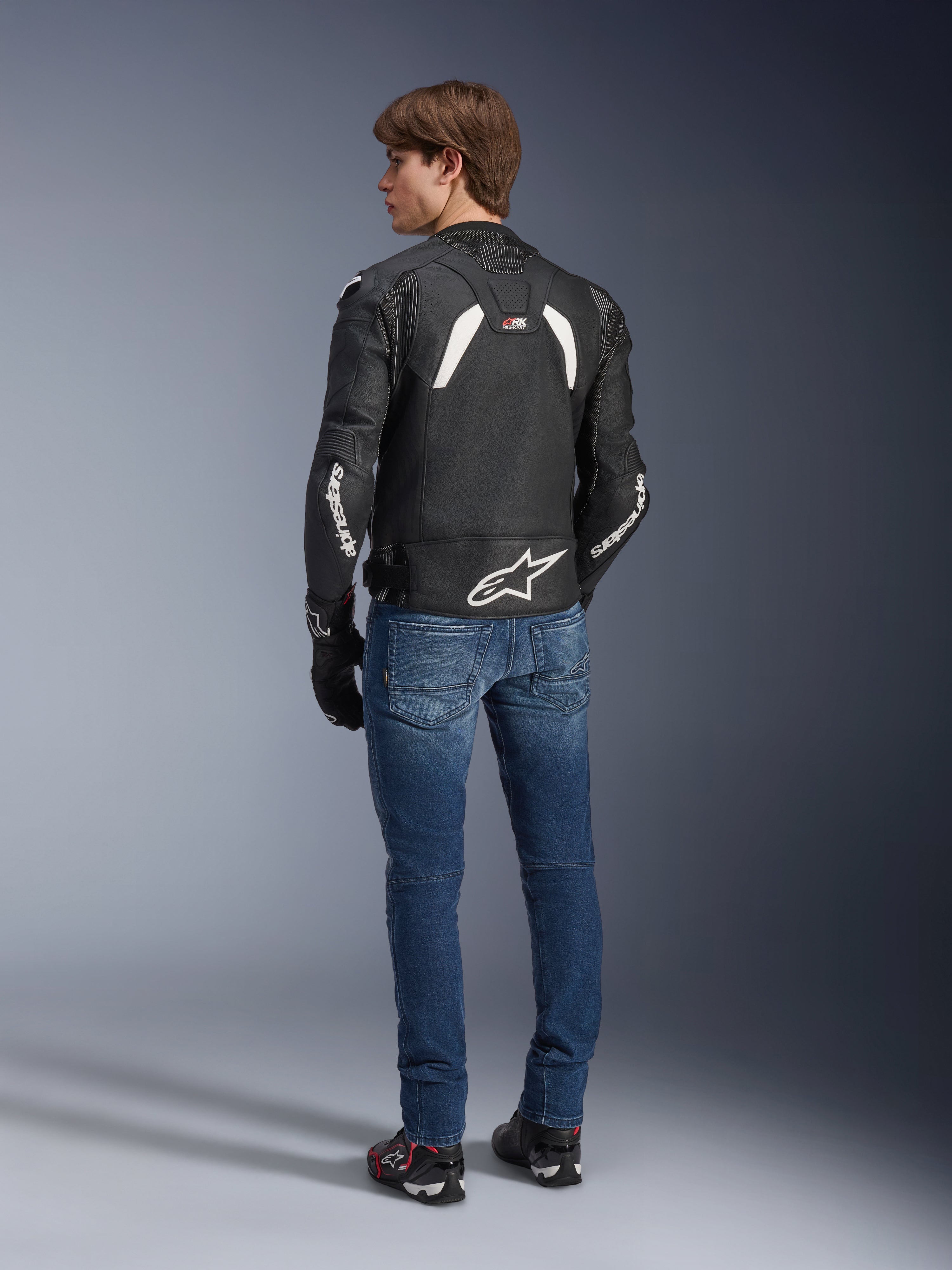 Chaqueta de cuero Alpinestars GP Plus R V4 Rideknit en negro y blanco, vista posterior en un modelo, con la marca Alpinestars en blanco en las mangas y un gran logotipo de estrella en la parte inferior de la espalda, usada con vaqueros azules y calzado de moto negro, equipamiento técnico de motociclismo.
