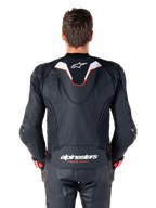Vista posterior de un piloto con la chaqueta de cuero Alpinestars Missile V3 Ignition, de cuero negro con detalles en blanco y rojo, con una joroba aerodinámica deportiva delgada con logo blanco y la marca 'alpinestars RACING AHEAD' en la parte inferior de la espalda, combinada con pantalones de carreras y guantes de cuero negro a juego.