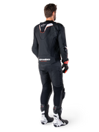 Vista trasera de un piloto con la chaqueta de cuero Alpinestars Missile V3 Ignition en color negro, que presenta una joroba aerodinámica deportiva delgada con detalles en blanco y rojo, logotipo de Alpinestars en la parte inferior de la espalda, combinada con pantalones de cuero negros a juego y botas de competición en blanco y negro; equipamiento profesional de conducción deportiva.