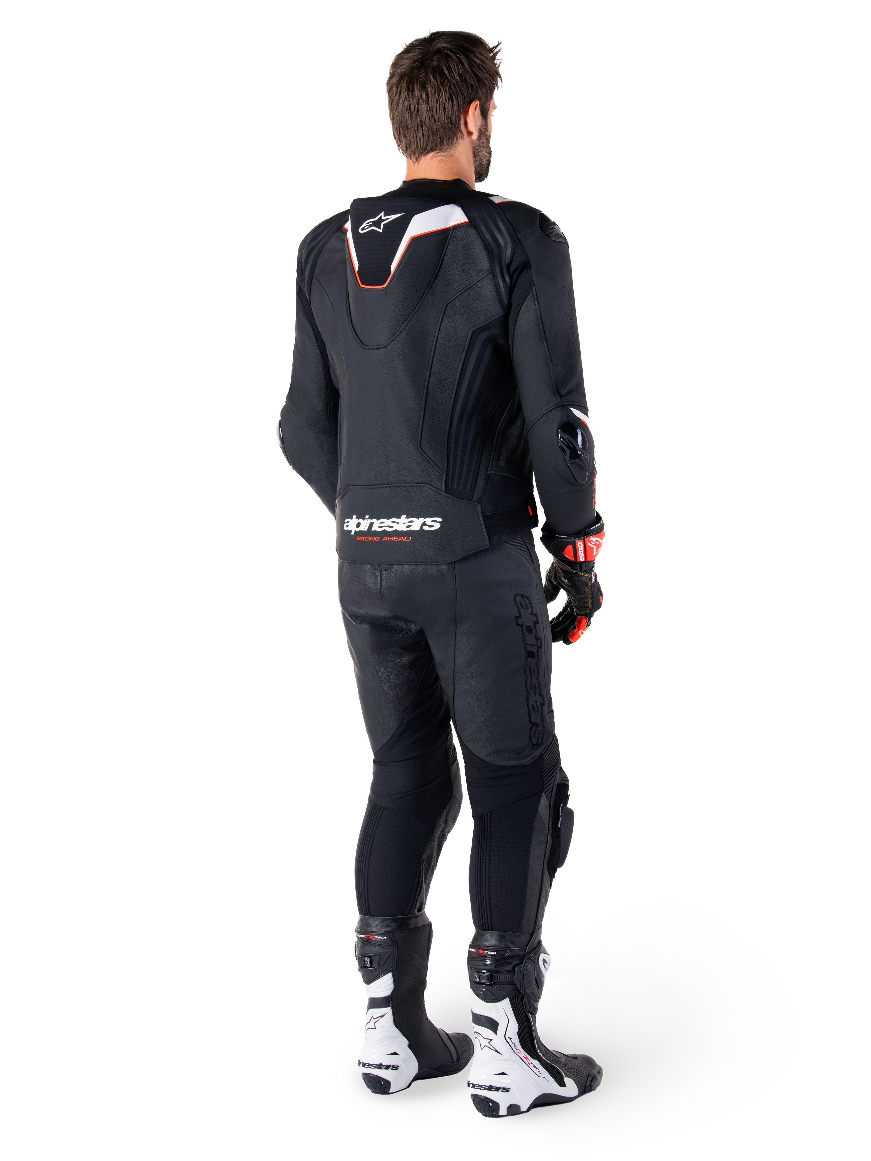 Vista trasera de un piloto con la chaqueta de cuero Alpinestars Missile V3 Ignition en color negro, que presenta una joroba aerodinámica deportiva delgada con detalles en blanco y rojo, logotipo de Alpinestars en la parte inferior de la espalda, combinada con pantalones de cuero negros a juego y botas de competición en blanco y negro; equipamiento profesional de conducción deportiva.