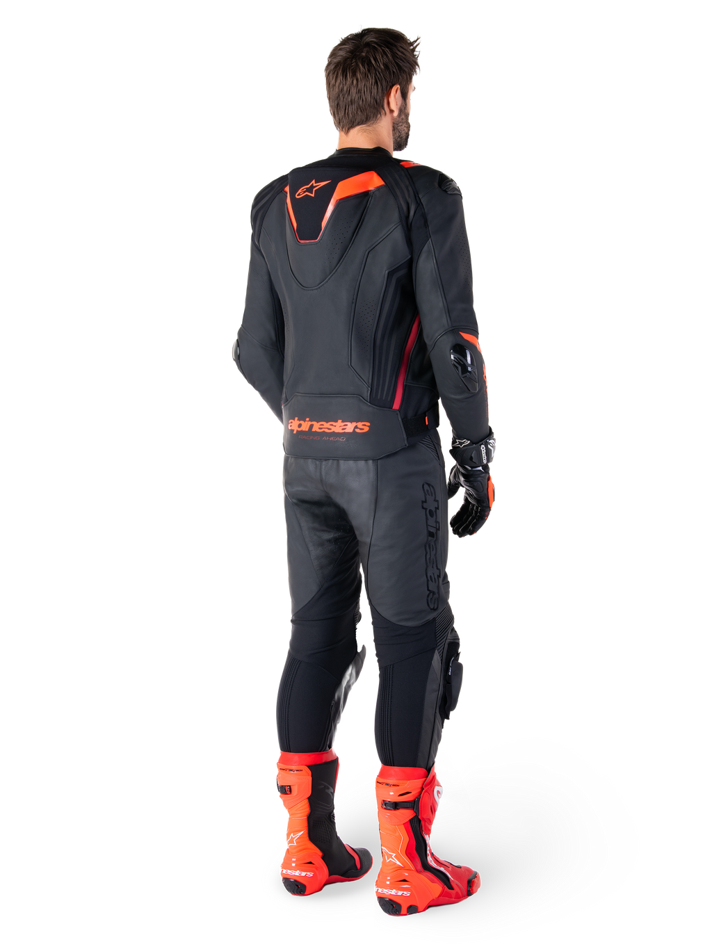 Alpinestars Missile V3 Ignition Leather Jacket, chaqueta de moto deportiva de competición, negro y rojo flúor, vista trasera de un piloto con equipación de carreras completa con pantalones de cuero, guantes y botas rojas a juego, con joroba aerodinámica deportiva y logotipo llamativo en la parte baja de la espalda.