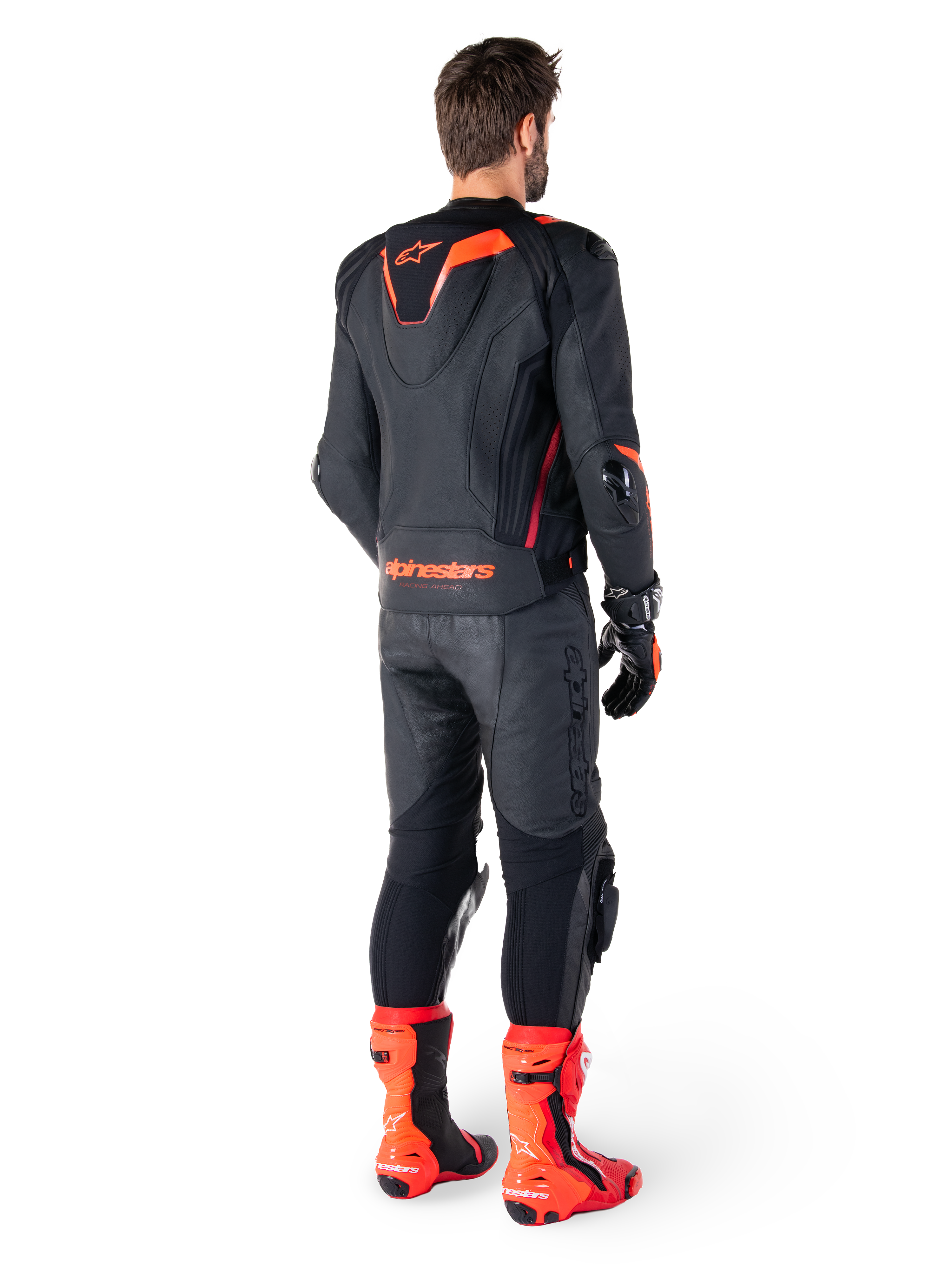 Alpinestars Missile V3 Ignition Leather Jacket, chaqueta de moto deportiva de competición, negro y rojo flúor, vista trasera de un piloto con equipación de carreras completa con pantalones de cuero, guantes y botas rojas a juego, con joroba aerodinámica deportiva y logotipo llamativo en la parte baja de la espalda.