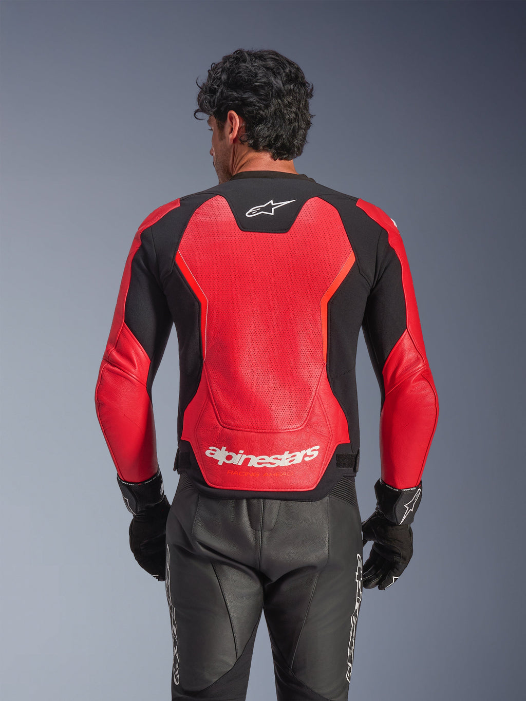 Chaqueta de cuero Alpinestars GP Force V2 Air en rojo y negro, vista trasera que muestra el chasis de cuero perforado para el flujo de aire, marca Alpinestars blanca en la parte baja de la espalda y paneles elásticos integrados, usada con pantalones y guantes de moto de cuero negro.