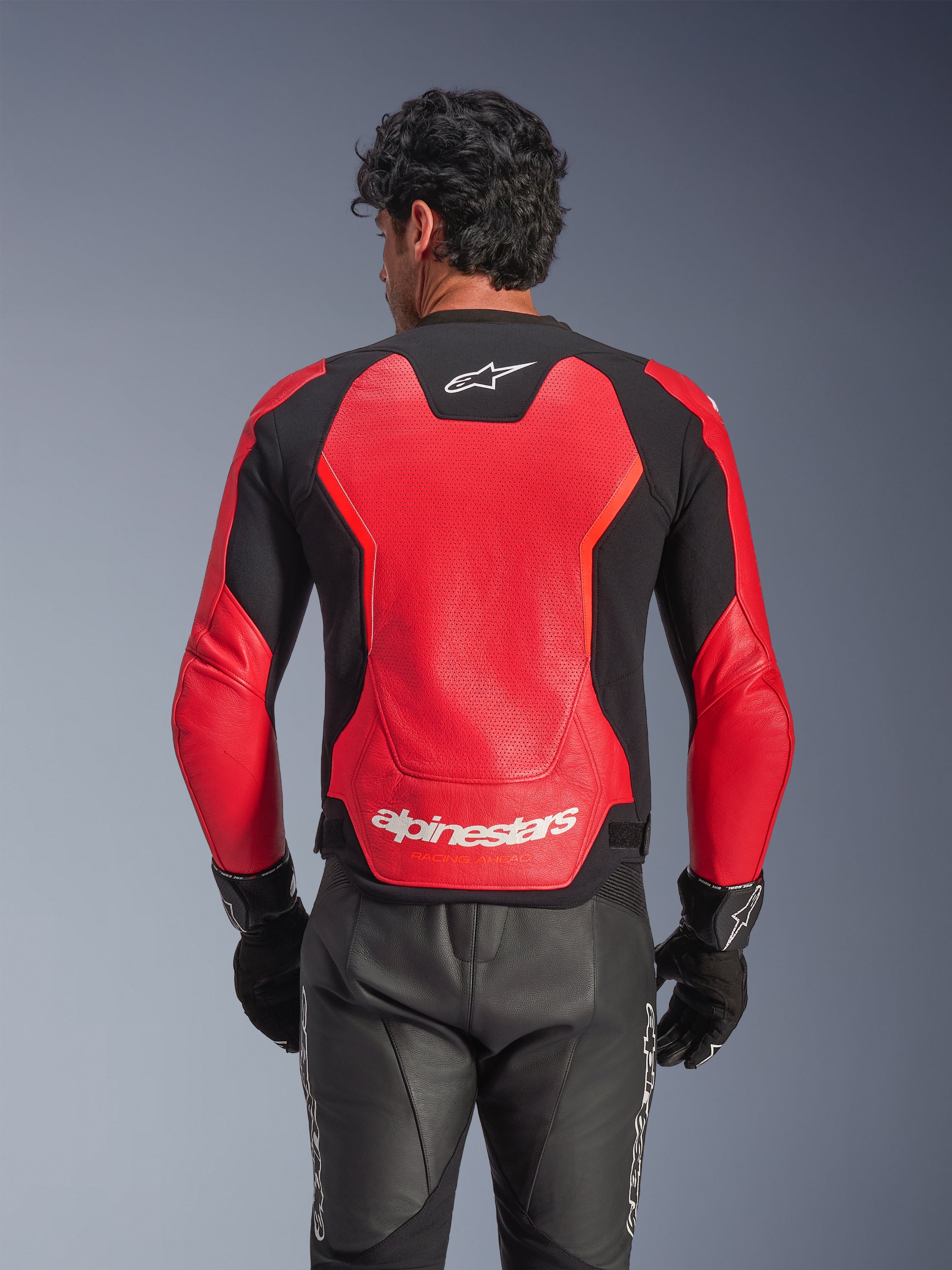 Chaqueta de cuero Alpinestars GP Force V2 Air en rojo y negro, vista trasera que muestra el chasis de cuero perforado para el flujo de aire, marca Alpinestars blanca en la parte baja de la espalda y paneles elásticos integrados, usada con pantalones y guantes de moto de cuero negro.