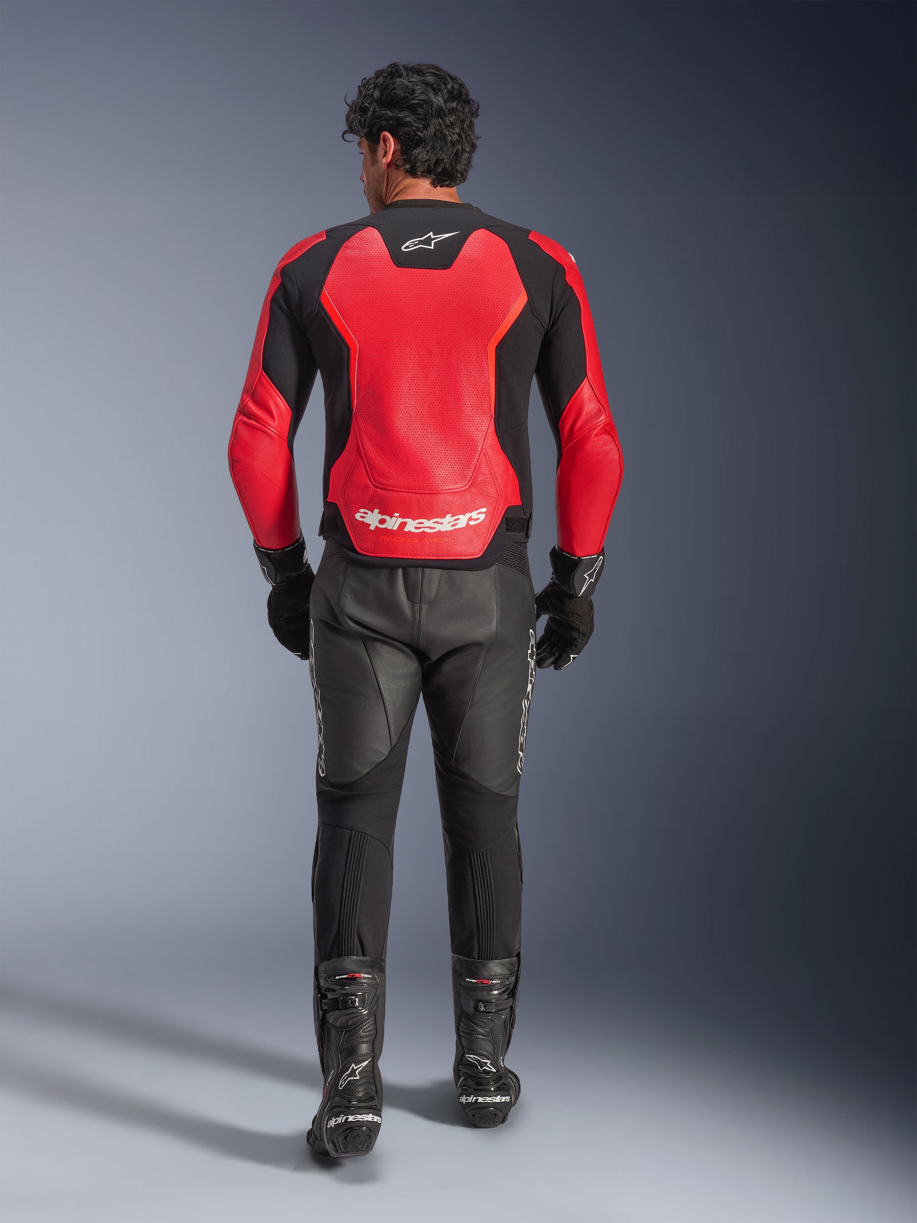 Vista trasera de un motorista con la chaqueta de cuero Alpinestars GP Force V2 Air en rojo y negro, con amplios paneles perforados y logotipo blanco de Alpinestars en la espalda, combinada con pantalones de cuero negros y botas de competición.
