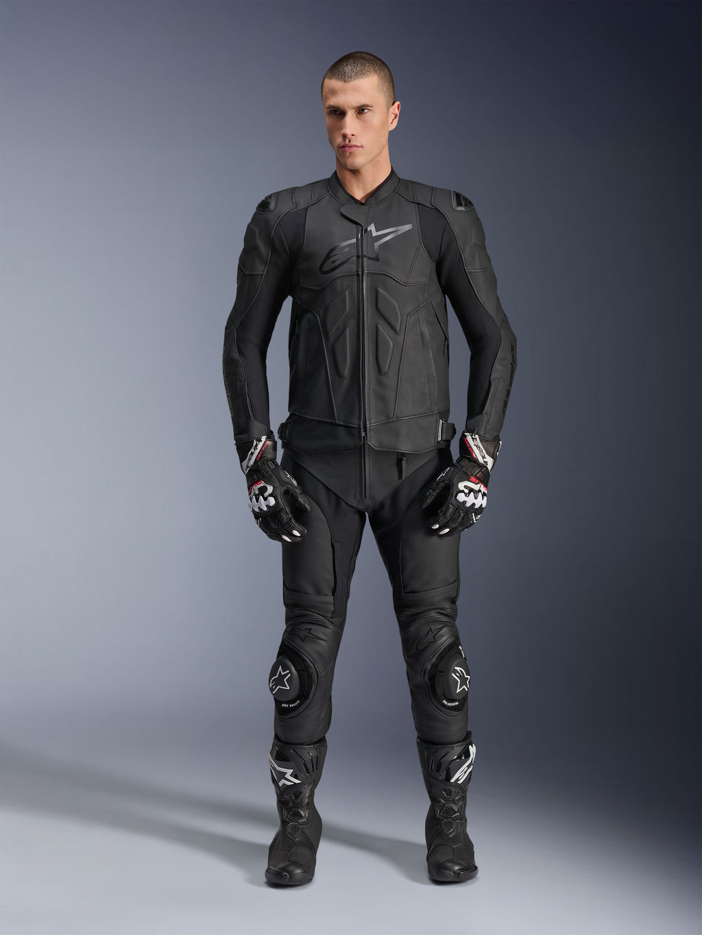 Chaqueta de cuero Alpinestars Dusk, negra, vestida por un modelo masculino como parte de un conjunto completo de inspiración racing con pantalones de cuero a juego, guantes y botas, diseño ergonómico elegante con protecciones en los hombros y logotipo en el pecho.