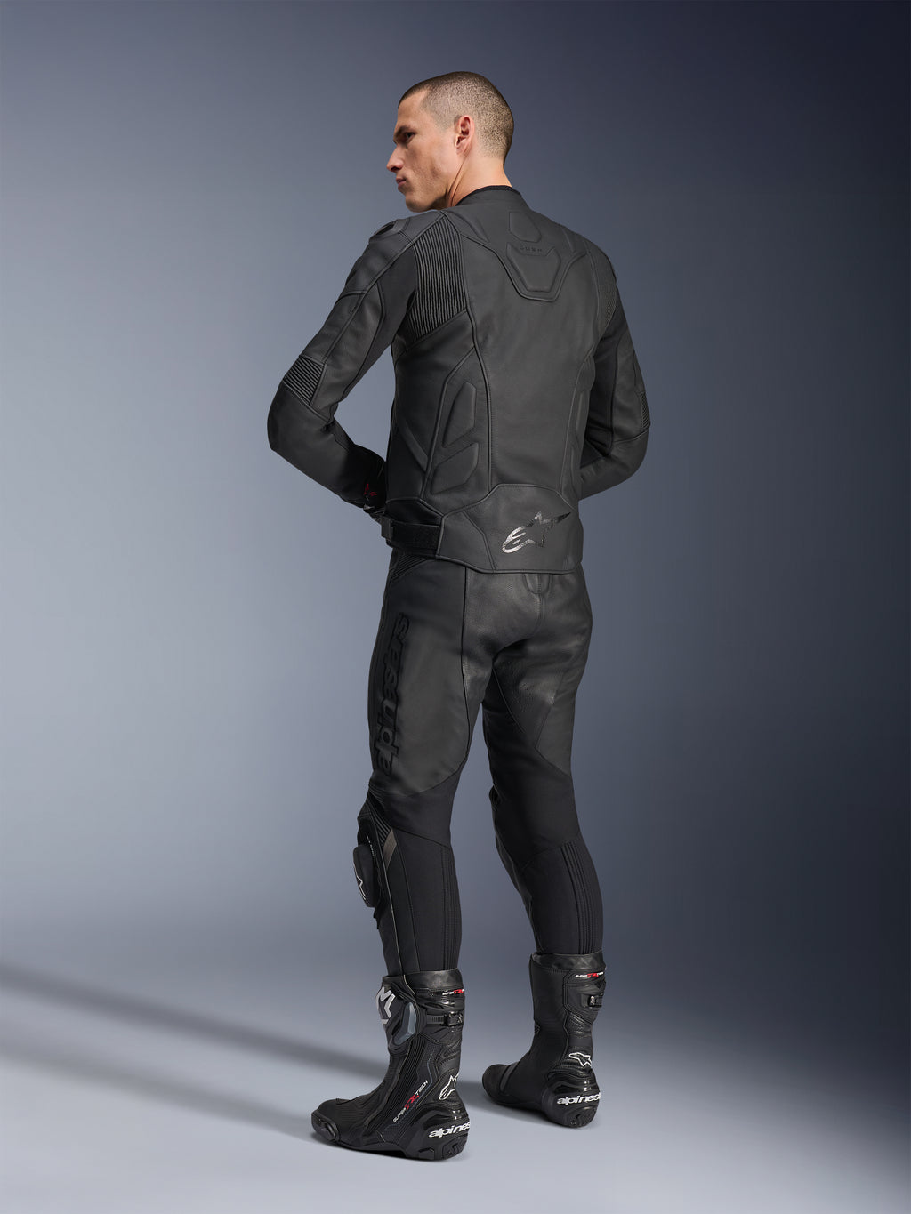 Chaqueta de cuero Alpinestars Dusk, negra, vista trasera de tres cuartos de un modelo con pantalones de moto de cuero a juego y botas de competición, con paneles elásticos de acordeón de cuero, logotipo de estrella Alpinestars plateado en la parte inferior de la espalda y marca DUSK en la parte superior, toma de estudio profesional