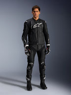 Modelo masculino con la chaqueta de cuero Alpinestars Dusk en negro y blanco, con logotipos blancos prominentes en el pecho y hombros, combinada con pantalones de moto de cuero negros a juego, guantes blancos y negros y botas negras, conjunto de competición deportiva completo para uso en carretera.