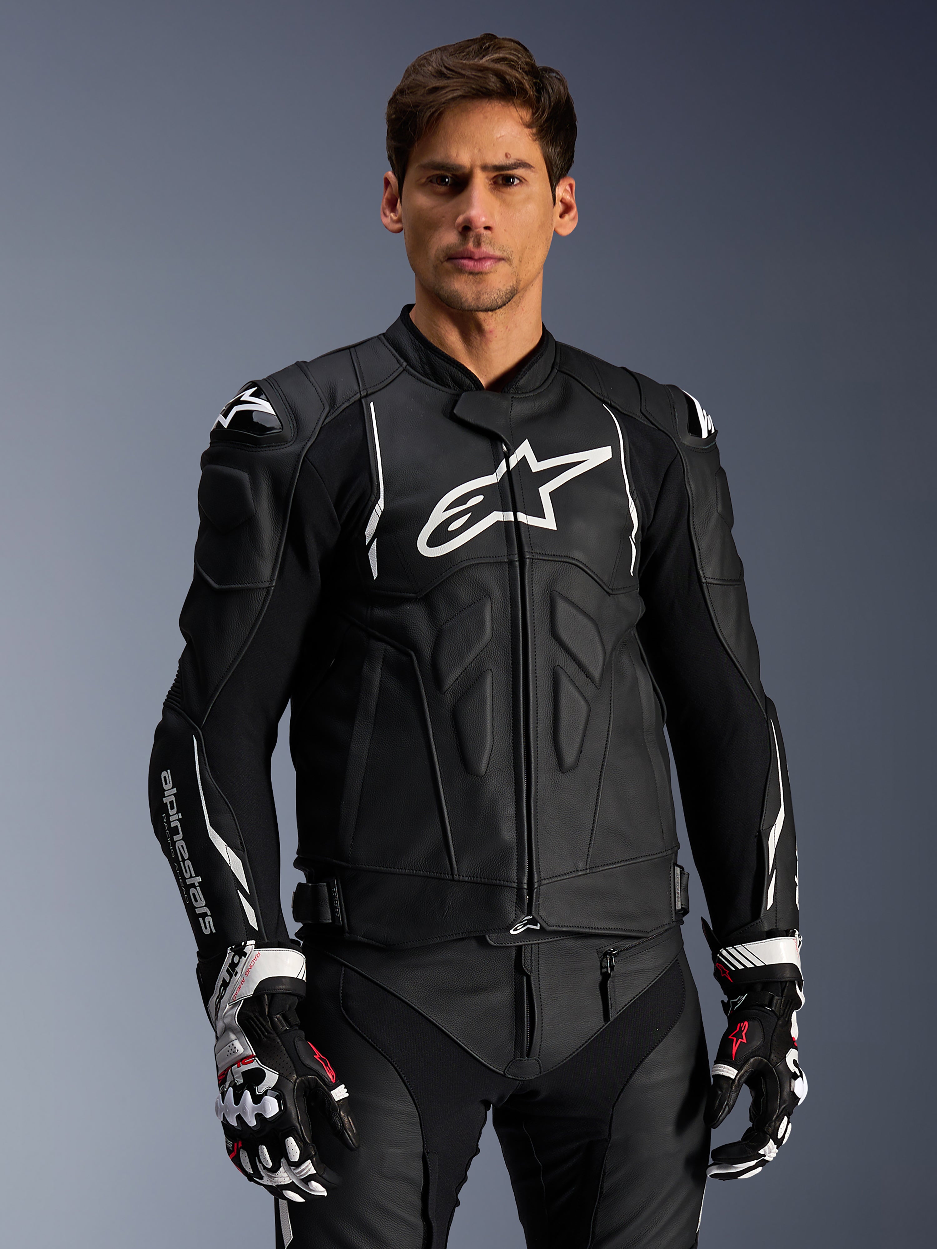 Chaqueta de cuero Alpinestars Dusk, negra y blanca, chaqueta de moto de cuero con áreas de protección acolchadas y logotipos blancos, combinada con guantes de carreras negros y blancos con detalles rojos, equipamiento profesional para conducción deportiva