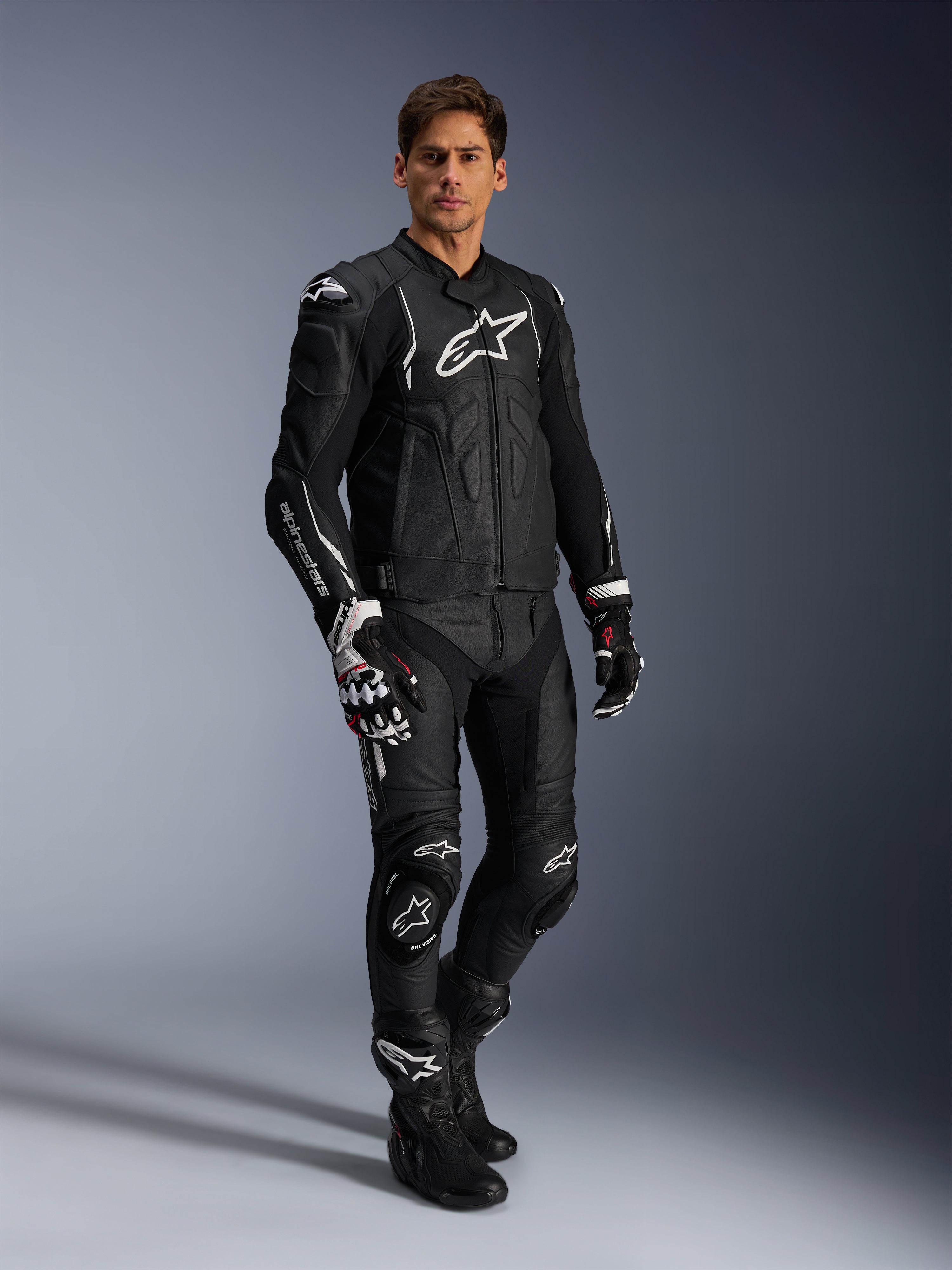 Piloto masculino con la chaqueta de cuero Alpinestars Dusk en negro y blanco, chaqueta de cuero de primera calidad con gran logotipo blanco en el pecho, combinada con pantalones de carreras de cuero a juego, guantes y botas, equipamiento deportivo de alto rendimiento completo.