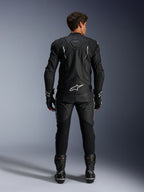 Piloto de moto visto desde atrás con la chaqueta de cuero Alpinestars Dusk y los pantalones de cuero Missile V3, traje de cuero negro con detalles blancos, la chaqueta presenta la marca 'Dusk' y el logotipo de Alpinestars en la espalda, combinado con botas y guantes de competición negros, conjunto de motociclismo coordinado para carretera.