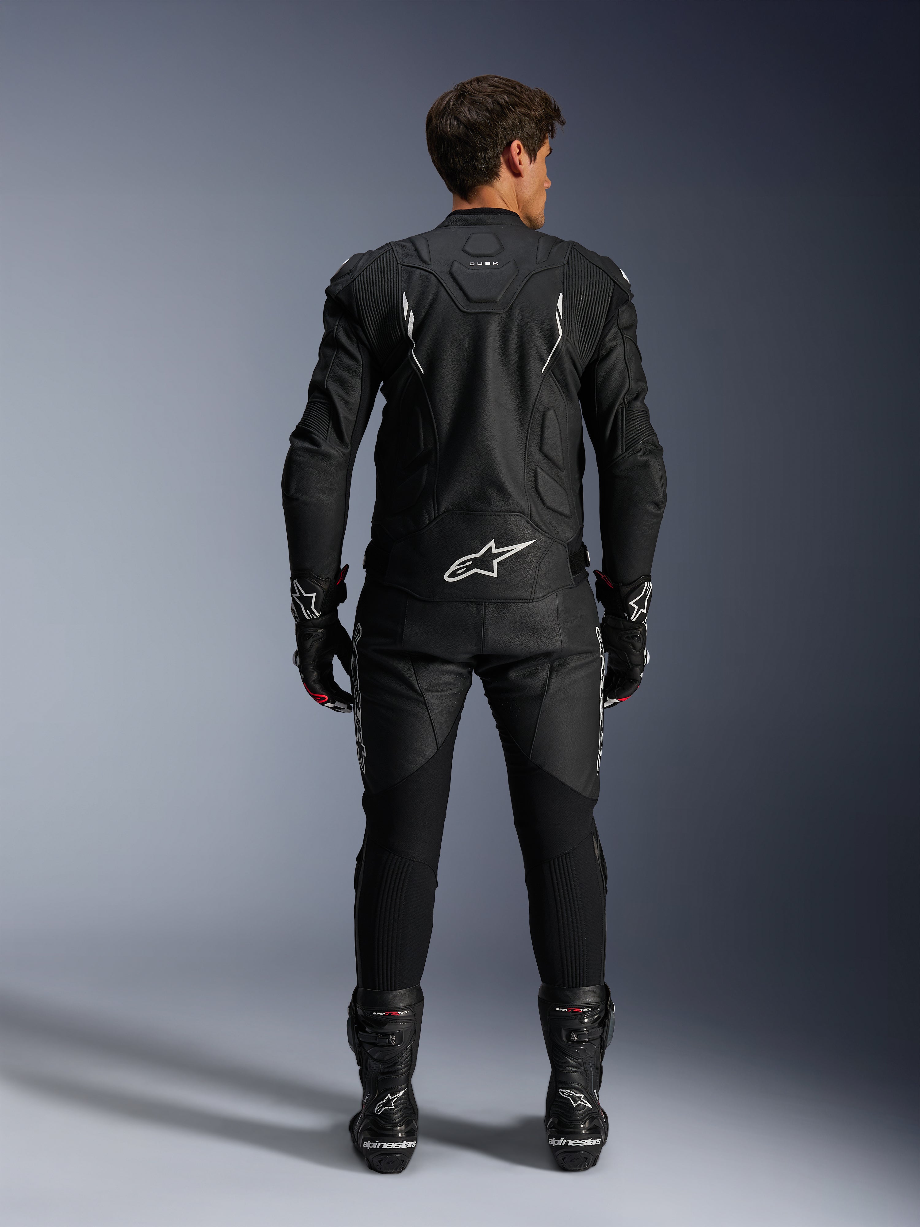 Piloto de moto visto desde atrás con la chaqueta de cuero Alpinestars Dusk y los pantalones de cuero Missile V3, traje de cuero negro con detalles blancos, la chaqueta presenta la marca 'Dusk' y el logotipo de Alpinestars en la espalda, combinado con botas y guantes de competición negros, conjunto de motociclismo coordinado para carretera.