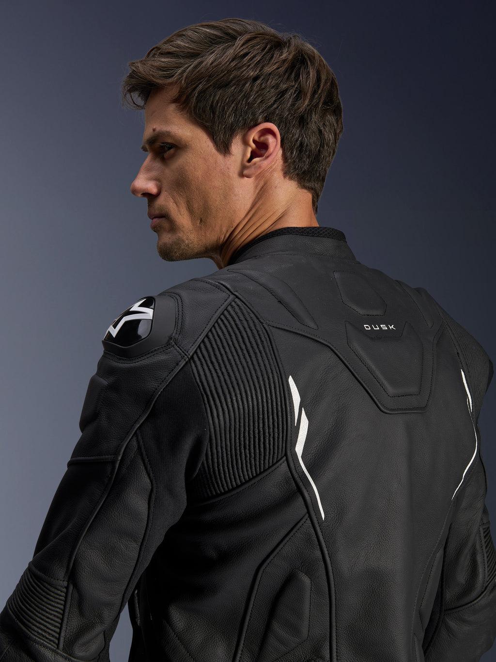 Chaqueta de cuero Alpinestars Dusk, negra con detalles en blanco, vista trasera sobre modelo, con protectores de hombros DFS con el logo de Alpinestars, paneles de cuero elástico tipo acordeón para movilidad y marca 'DUSK' en la parte superior de la espalda, chaqueta de cuero bovino premium para conducción en carretera y deportiva