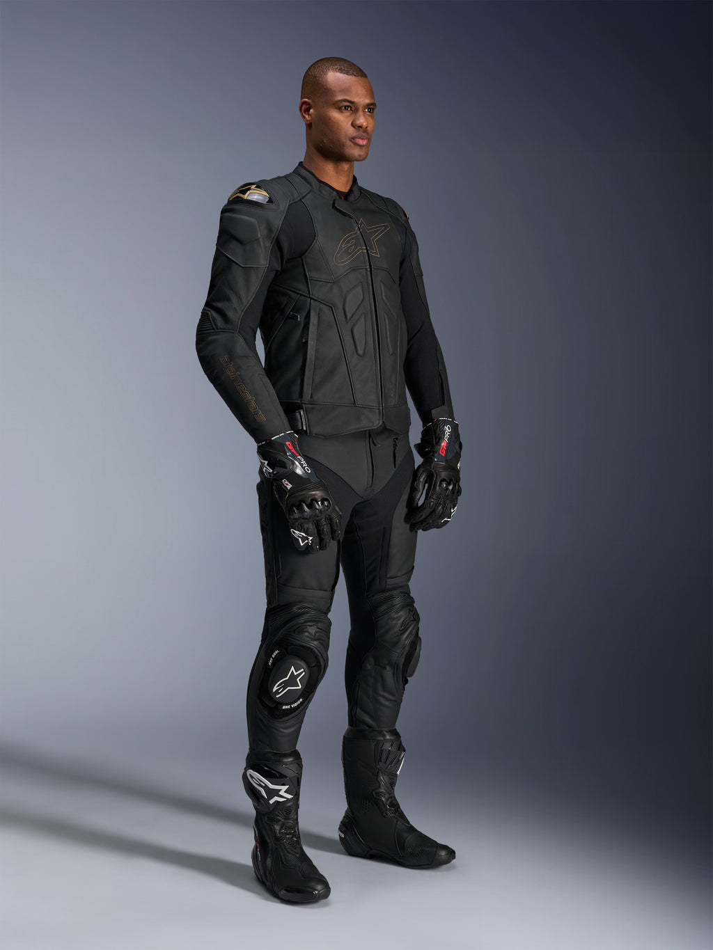 Modelo masculino con pantalones de cuero Alpinestars Missile V3 y chaqueta de cuero negra a juego con detalles dorados, guantes de competición GP Pro negros y botas de carreras negras, equipo completo de motociclismo para carretera, foto de estudio