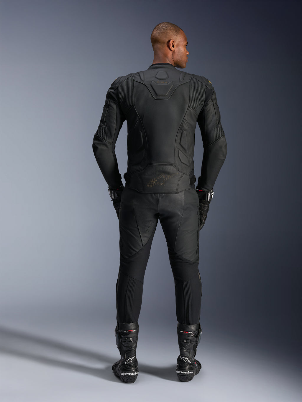 Chaqueta de cuero Alpinestars Dusk, Black Laser, vista trasera en un piloto, usada con pantalones de moto de cuero a juego, guantes de carreras y botas, con construcción ergonómica 3D de paneles múltiples y paneles de cuero protectores.
