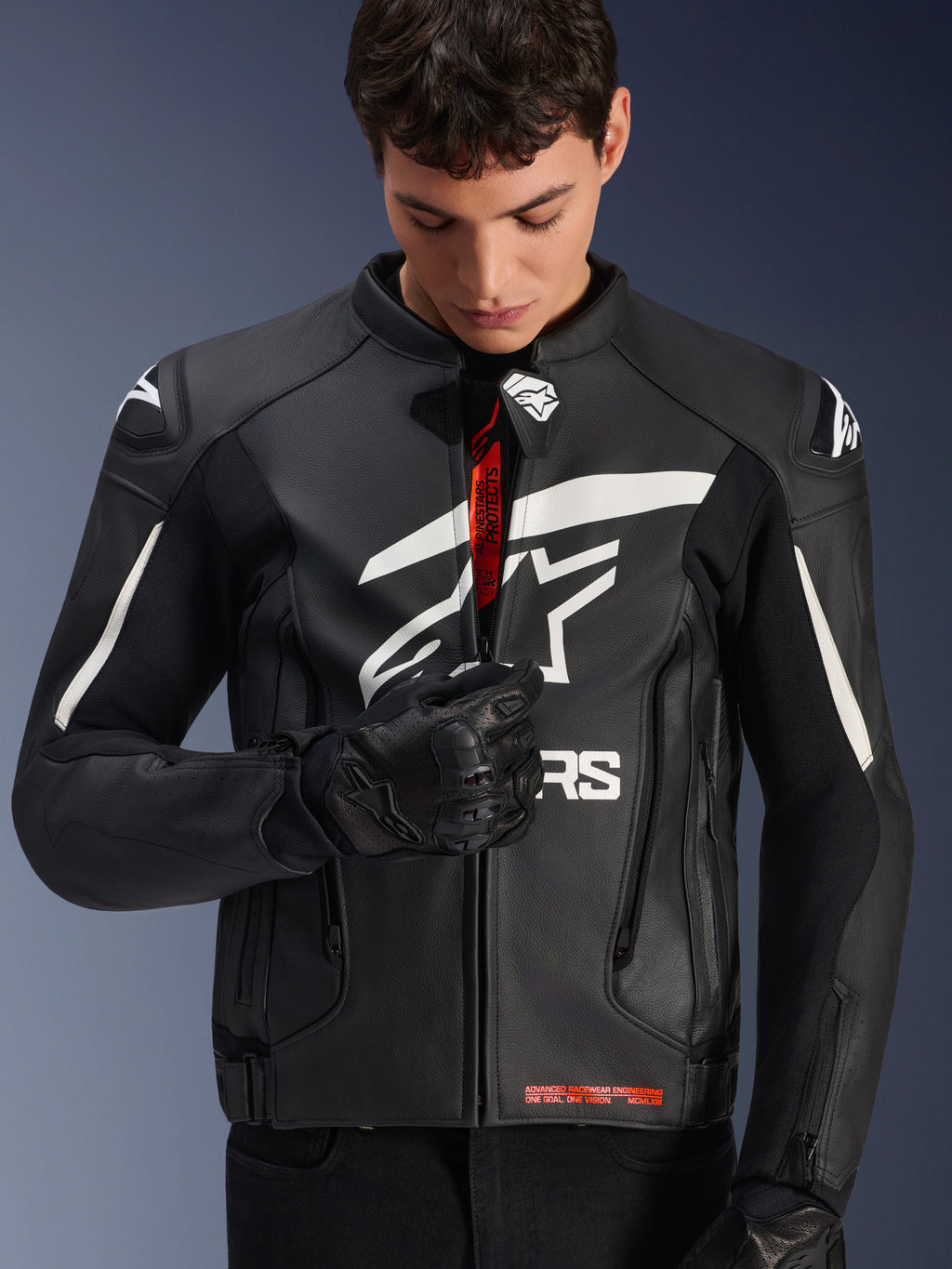 Chaqueta de cuero Alpinestars GP Plus V4, negro y blanco, modelo masculino subiendo la cremallera frontal para mostrar el branding rojo interior, con logotipos Alpinestars blancos y llamativos en el pecho y los hombros, combinada con guantes de moto de cuero negro, equipamiento de cuero para conducción deportiva