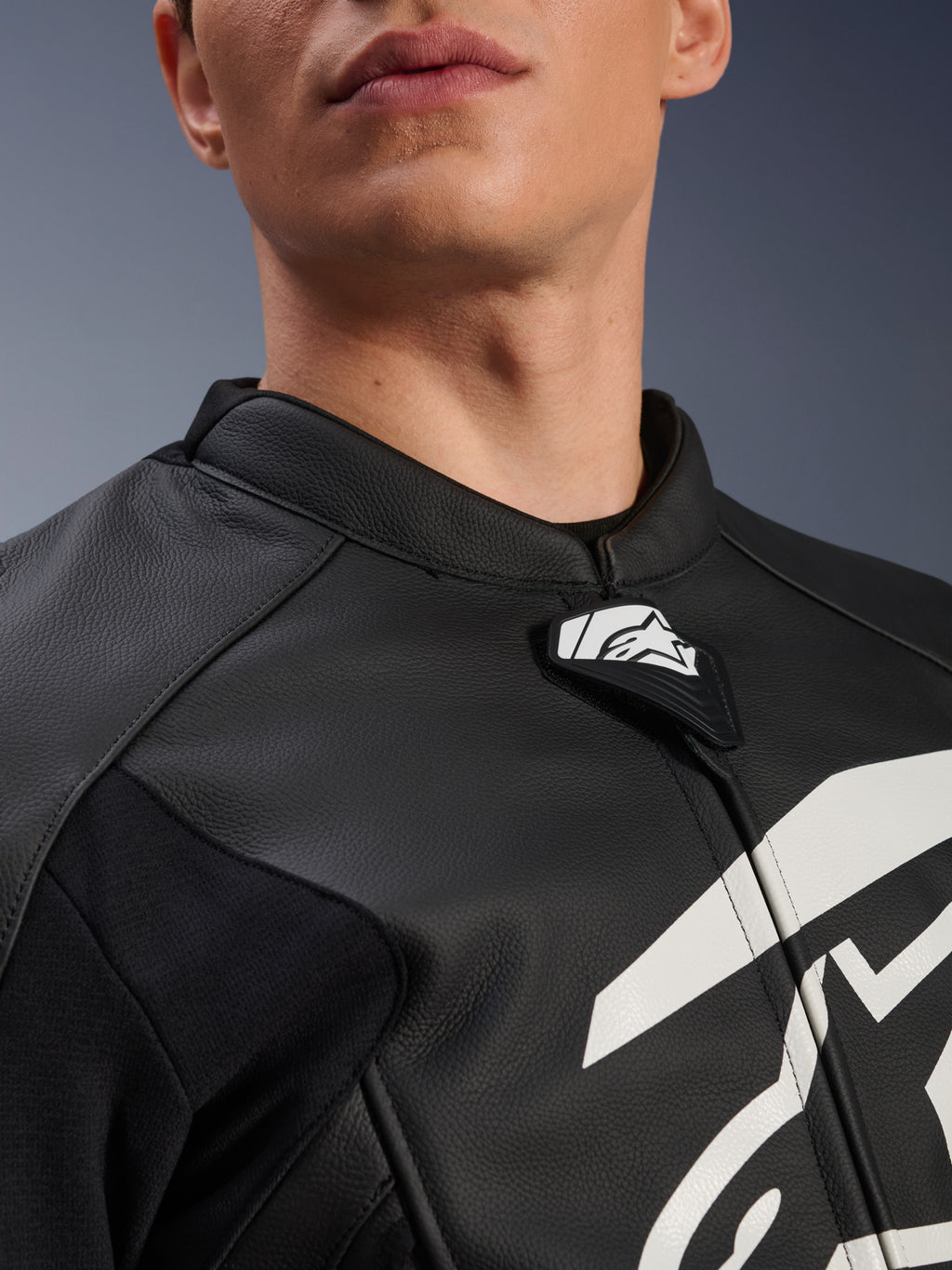 Alpinestars GP Plus V4 Leather Jacket, chaqueta deportiva de moto, negra y blanca, primer plano del cuello de cuero texturizado y la lengüeta con el logo de Alpinestars, gráfico blanco en el pecho, diseño derivado de la competición