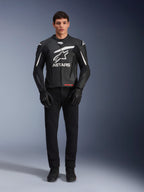 Modelo masculino con la chaqueta de cuero Alpinestars GP Plus V4, en color negro y blanco, con el llamativo logotipo blanco de Astars en el pecho y detalles blancos en los hombros, combinada con guantes y pantalones de moto negros, equipamiento de motociclismo deportivo de diseño estilizado