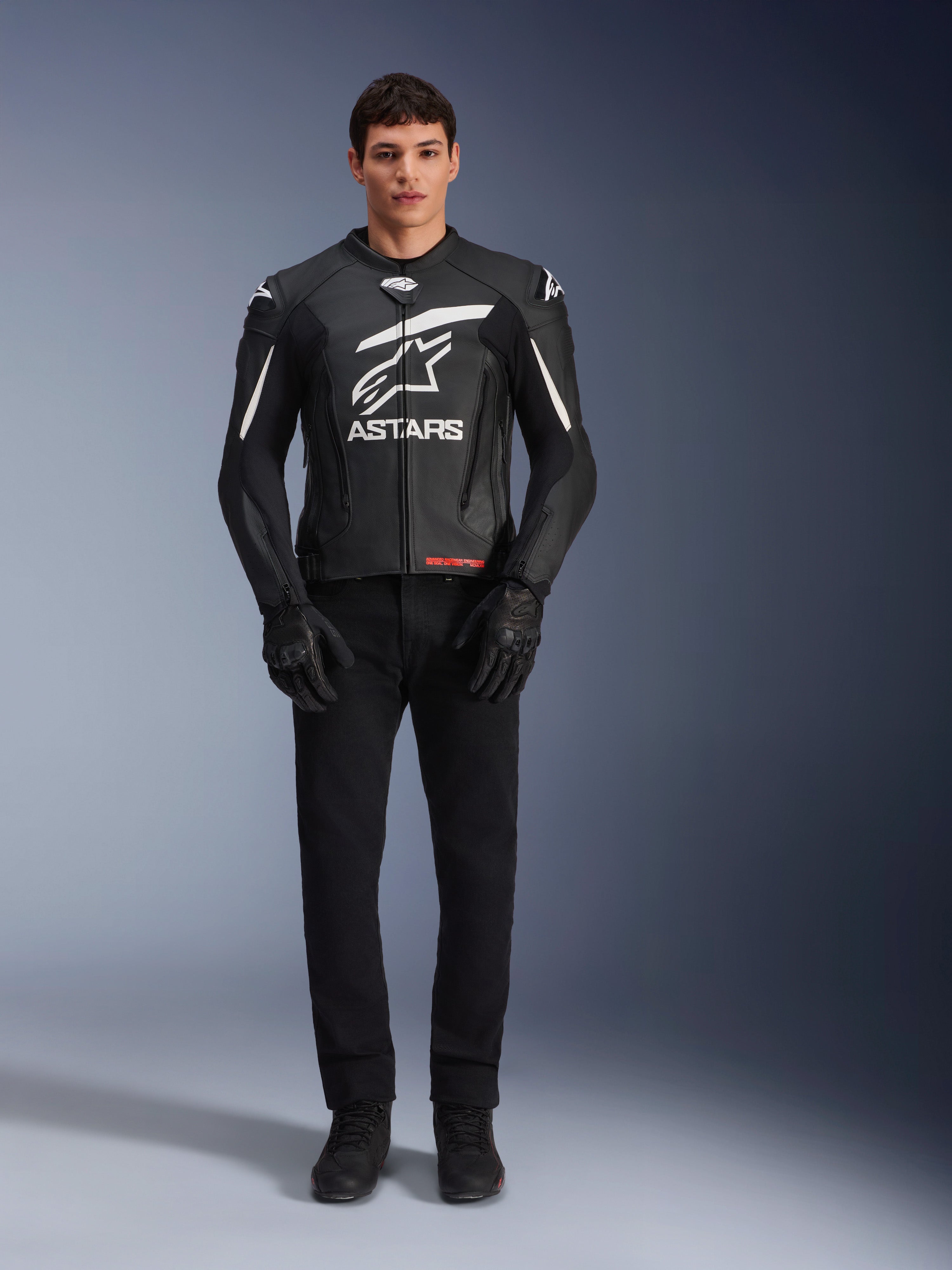 Modelo masculino con la chaqueta de cuero Alpinestars GP Plus V4, en color negro y blanco, con el llamativo logotipo blanco de Astars en el pecho y detalles blancos en los hombros, combinada con guantes y pantalones de moto negros, equipamiento de motociclismo deportivo de diseño estilizado