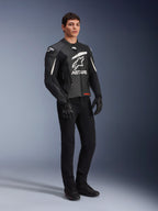 Chaqueta de cuero Alpinestars GP Plus V4, chaqueta deportiva de moto, en negro y blanco, lucida por un modelo masculino con pantalones de montar y guantes negros, con un gran logotipo ASTARS blanco en el pecho y deslizadores externos de hombro DFS, diseño profesional derivado de la competición