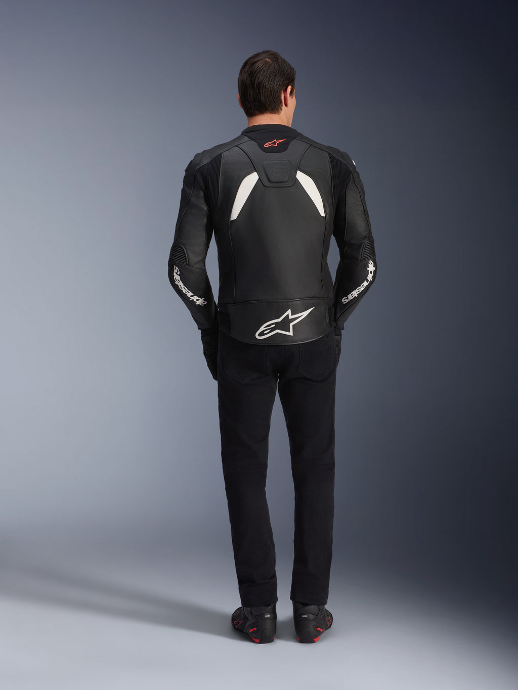 Modelo visto desde atrás vistiendo la chaqueta de cuero Alpinestars GP Plus V4 en negro y blanco, combinada con pantalones negros y botas de moto, con paneles blancos en los hombros y el logotipo de Alpinestars en las mangas y la parte inferior de la espalda.