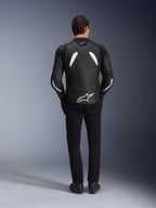 Modelo visto desde atrás vistiendo la chaqueta de cuero Alpinestars GP Plus V4 en negro y blanco, combinada con pantalones negros y botas de moto, con paneles blancos en los hombros y el logotipo de Alpinestars en las mangas y la parte inferior de la espalda.