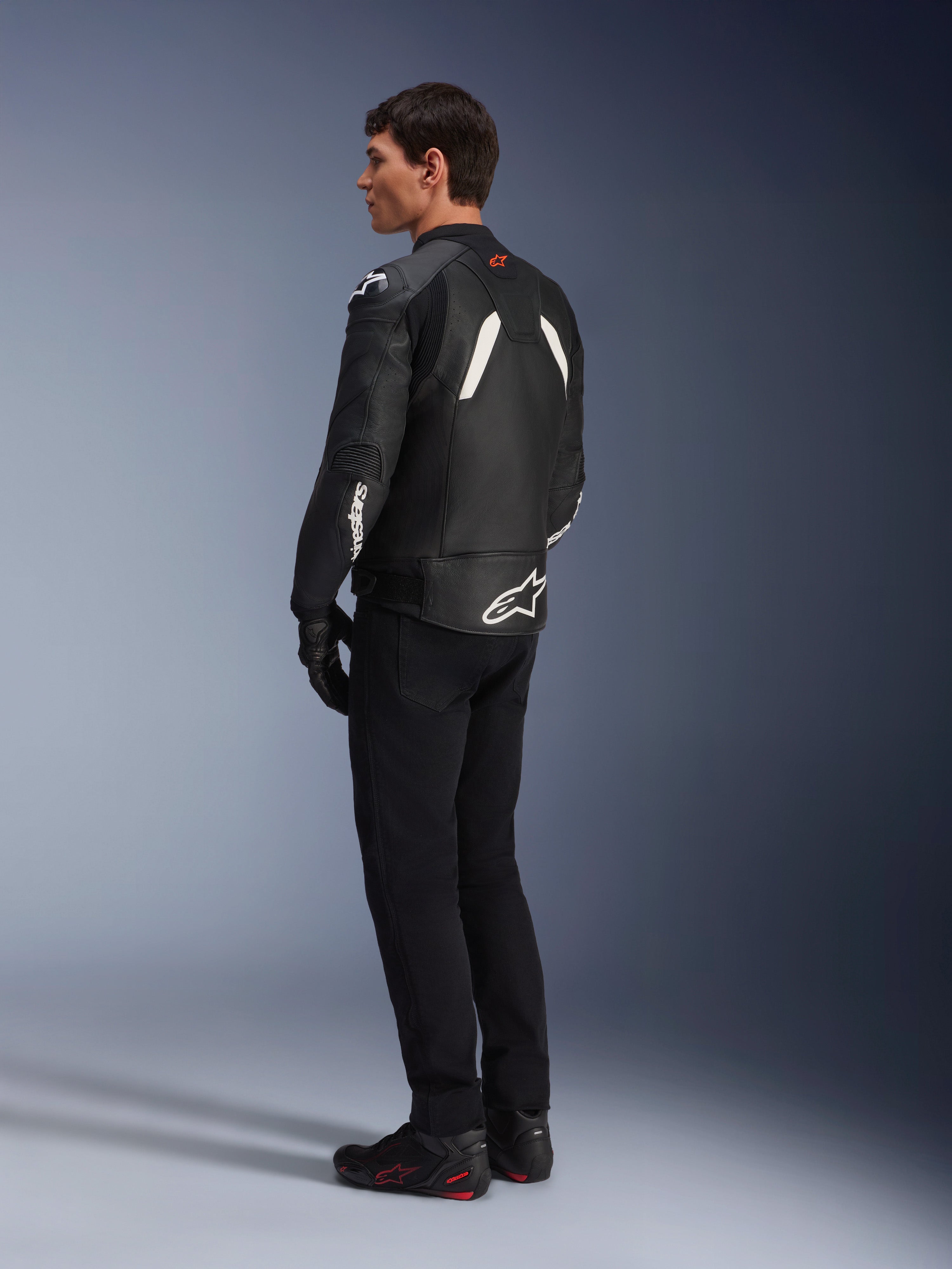 Chaqueta de cuero Alpinestars GP Plus V4, chaqueta deportiva de cuero, negro y blanco, vista de perfil posterior en un modelo, con logotipos blancos de Alpinestars en la parte inferior de la espalda y la manga, combinada con pantalones negros y botas técnicas para un look de rendimiento deportivo.