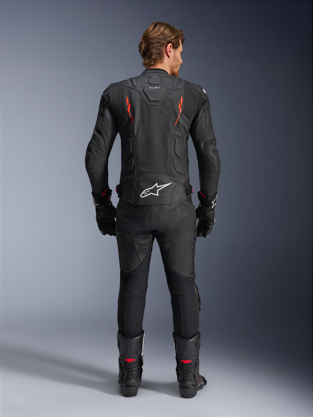 Chaqueta de cuero Alpinestars Dusk Airflow, negro y rojo flúor, vista trasera con perforaciones extensas y un gran logotipo blanco en la parte baja de la espalda, combinada con equipamiento de carreras de cuero a juego que incluye pantalones, botas y guantes