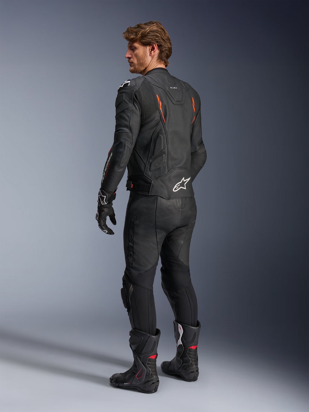 Chaqueta de cuero Alpinestars Dusk Airflow, negro con detalles en rojo flúor, vista trasera de un piloto con equipamiento de cuero completo incluyendo pantalones negros, guantes y botas de competición, con cuero perforado y logotipos blancos de Alpinestars