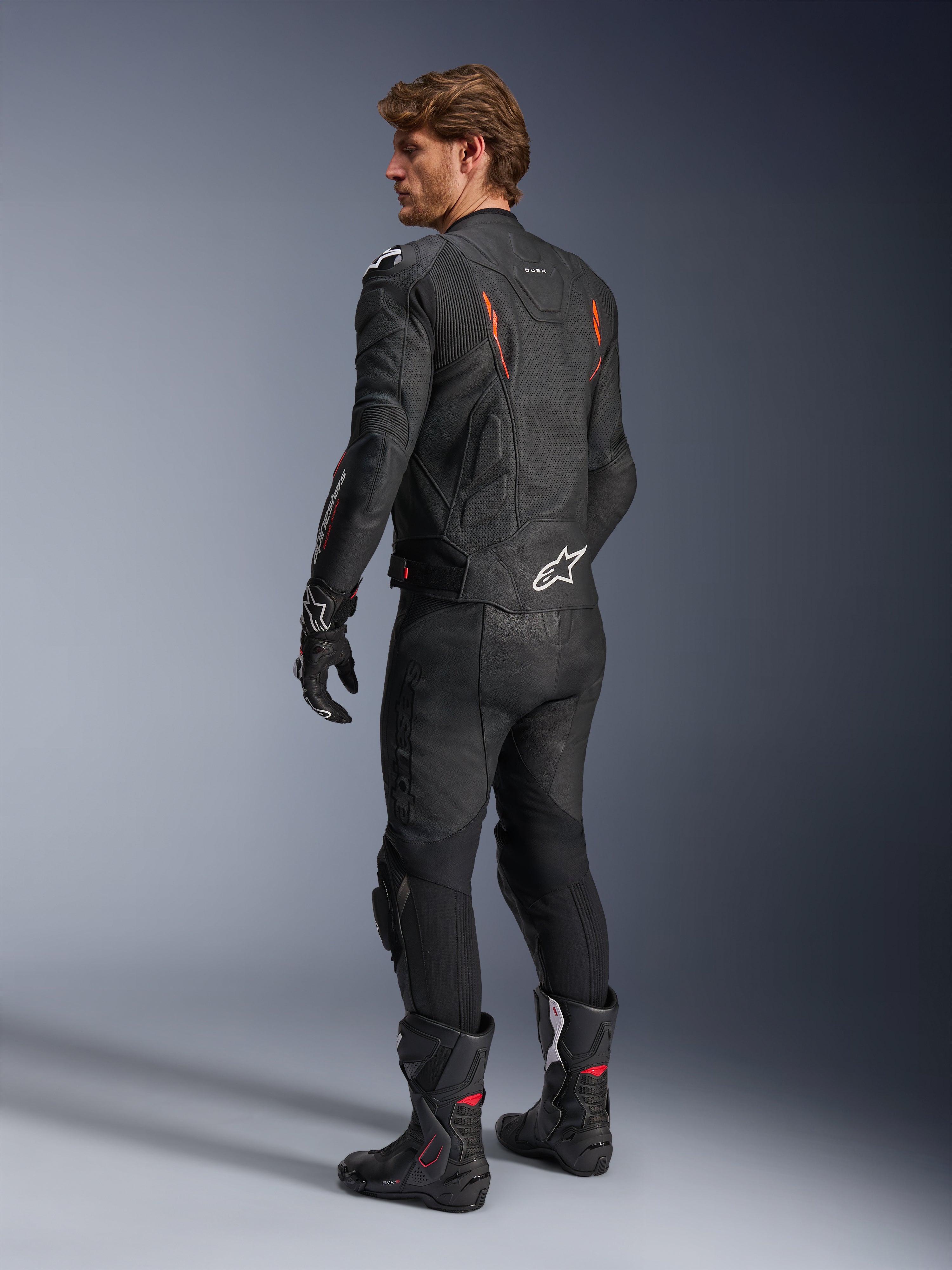 Chaqueta de cuero Alpinestars Dusk Airflow, negro con detalles en rojo flúor, vista trasera de un piloto con equipamiento de cuero completo incluyendo pantalones negros, guantes y botas de competición, con cuero perforado y logotipos blancos de Alpinestars