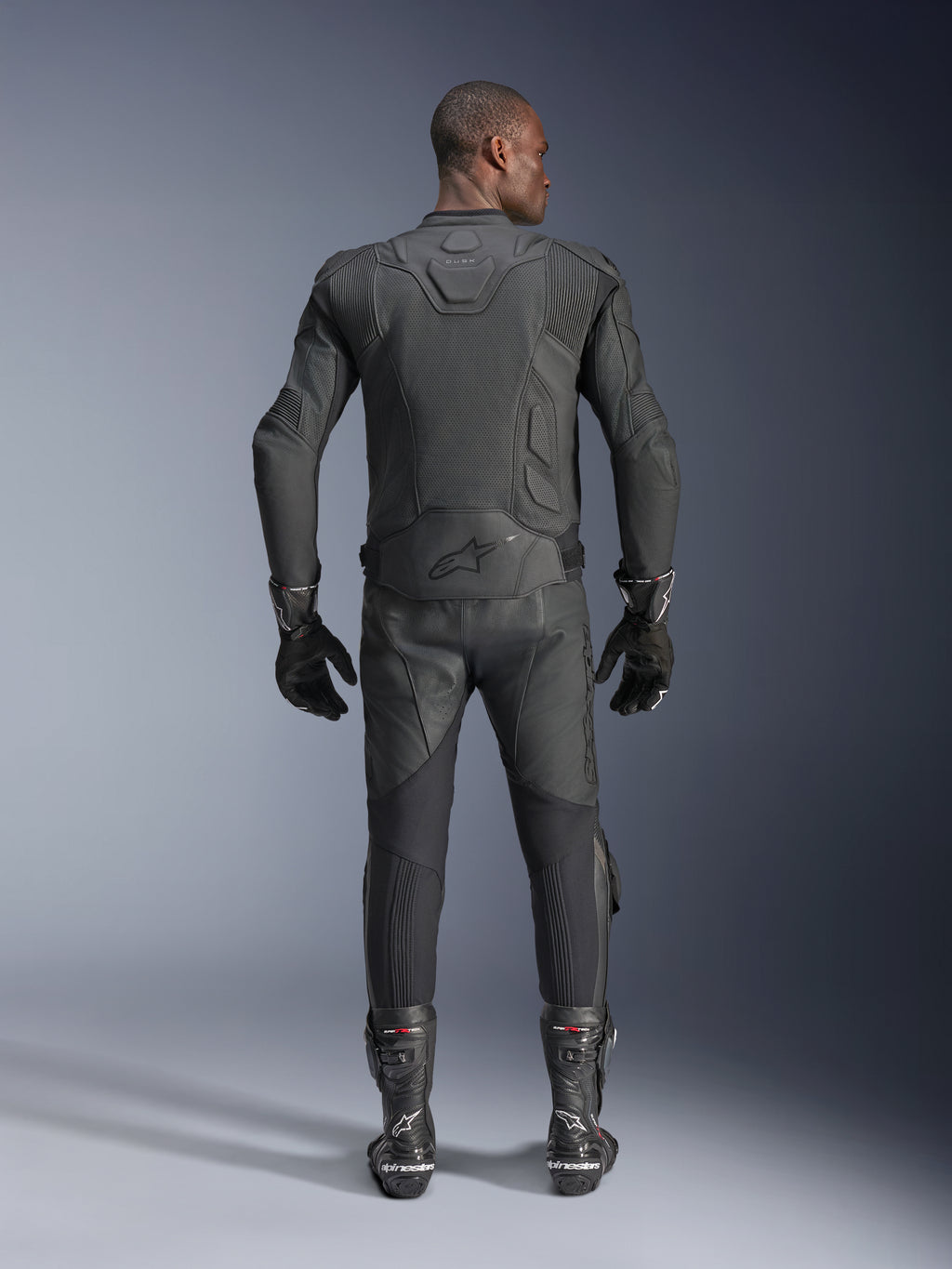 Modelo masculino visto desde atrás luciendo la chaqueta de cuero Alpinestars Dusk Airflow en color negro, combinada con pantalones de carreras de cuero negro, guantes y botas a juego, con perforaciones extensas para ventilación y el logotipo de Alpinestars en la parte inferior de la espalda.