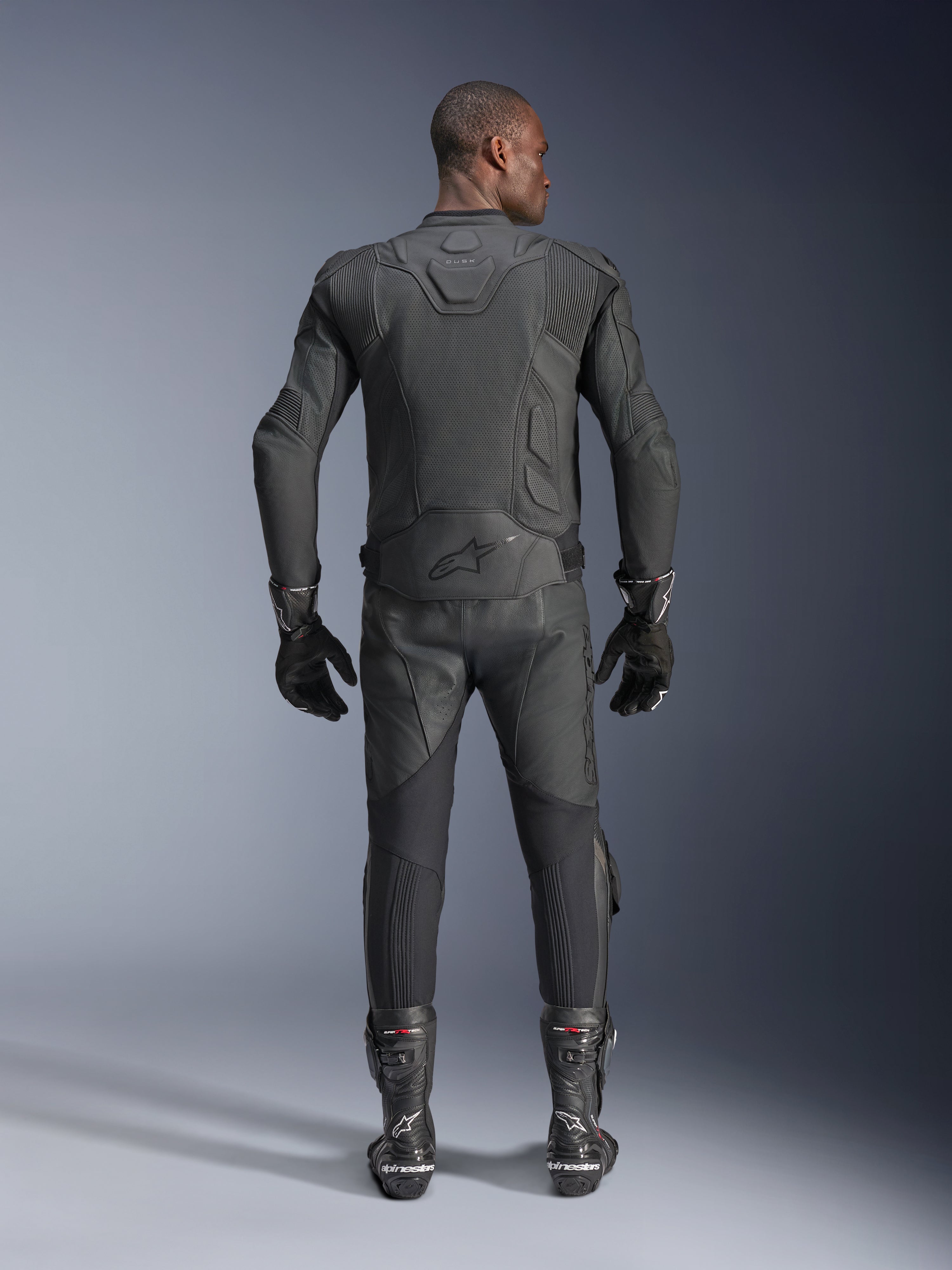Modelo masculino visto desde atrás luciendo la chaqueta de cuero Alpinestars Dusk Airflow en color negro, combinada con pantalones de carreras de cuero negro, guantes y botas a juego, con perforaciones extensas para ventilación y el logotipo de Alpinestars en la parte inferior de la espalda.