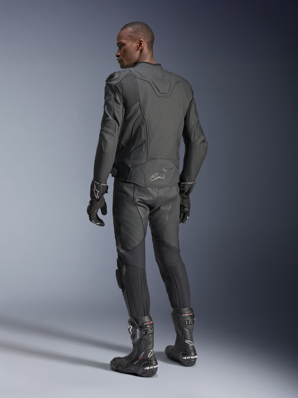 Piloto masculino de espaldas vistiendo la chaqueta de cuero Alpinestars Dusk Airflow en color Black Black, combinada con pantalones de cuero, guantes y botas a juego, destacando las amplias perforaciones en la espalda y el ajuste deportivo de alto rendimiento.