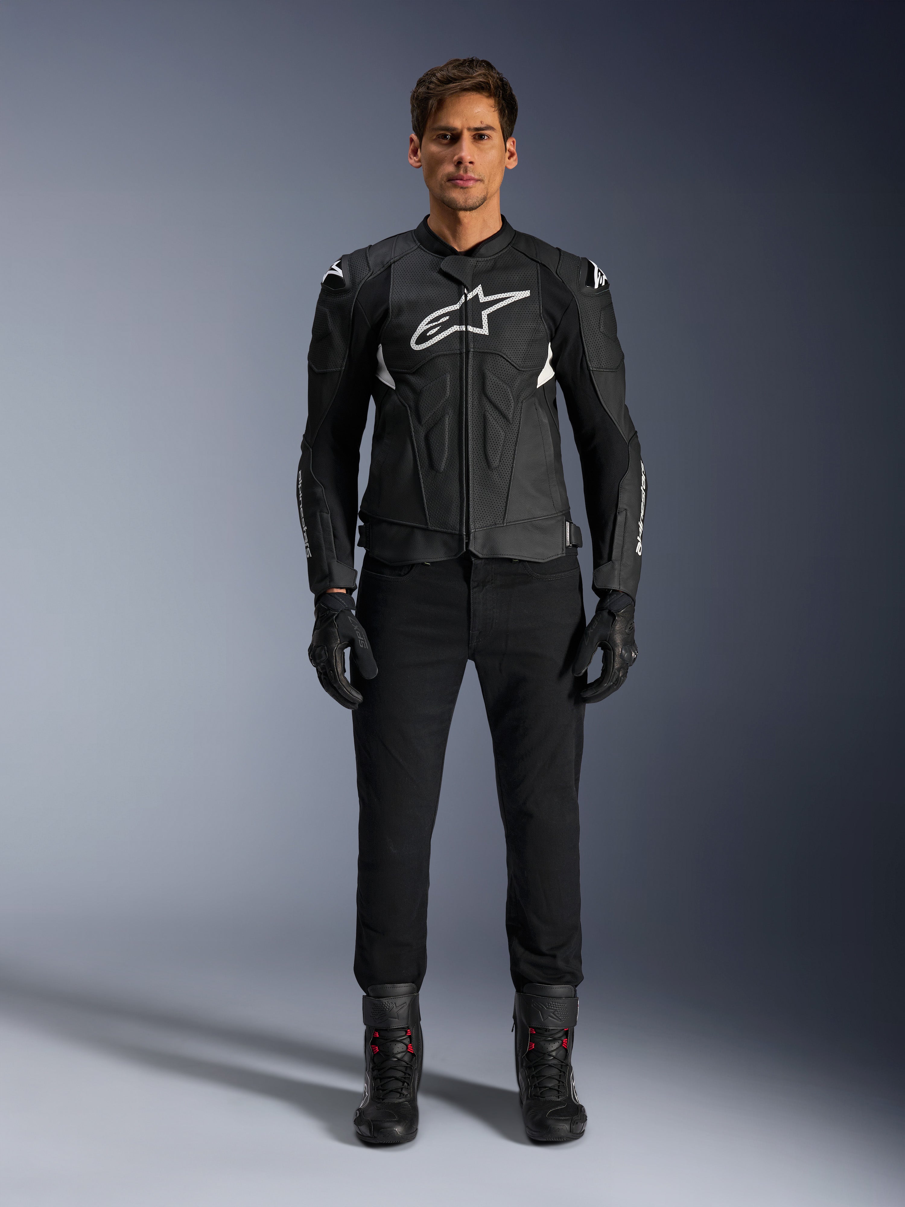 Chaqueta de cuero Alpinestars Dusk Airflow, negra y blanca, chaqueta de cuero para moto con paneles perforados para el flujo de aire, logotipo Astars blanco en el pecho, usada por un piloto masculino con pantalones negros, guantes y botas de conducción, toma de estudio profesional para equipamiento de moto deportiva.
