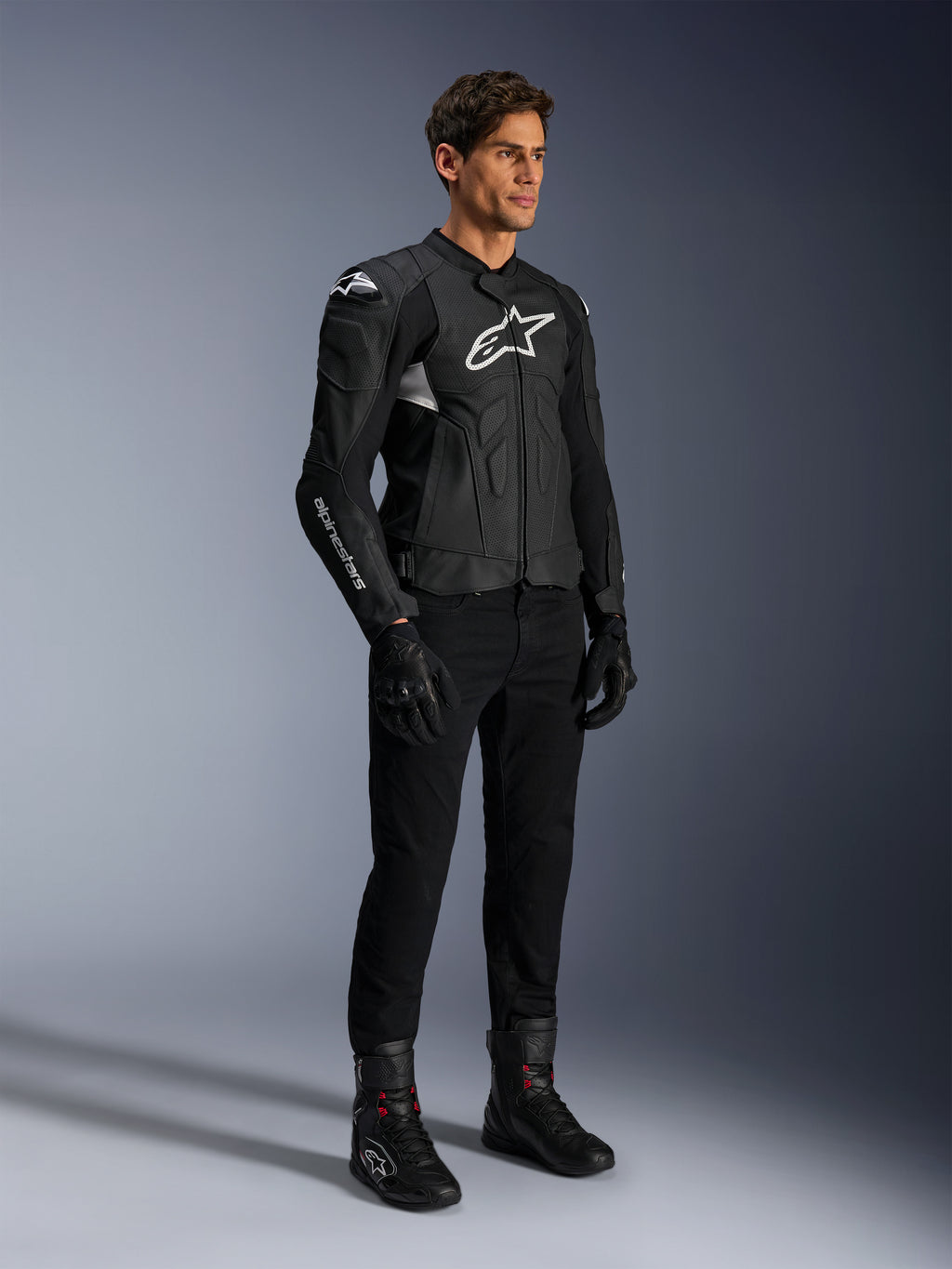 Chaqueta de cuero Alpinestars Dusk Airflow, negra y blanca, modelada con pantalones de moto, guantes y botas negros, con paneles de cuero perforado y logotipos blancos, estilo deportivo de competición en carretera.