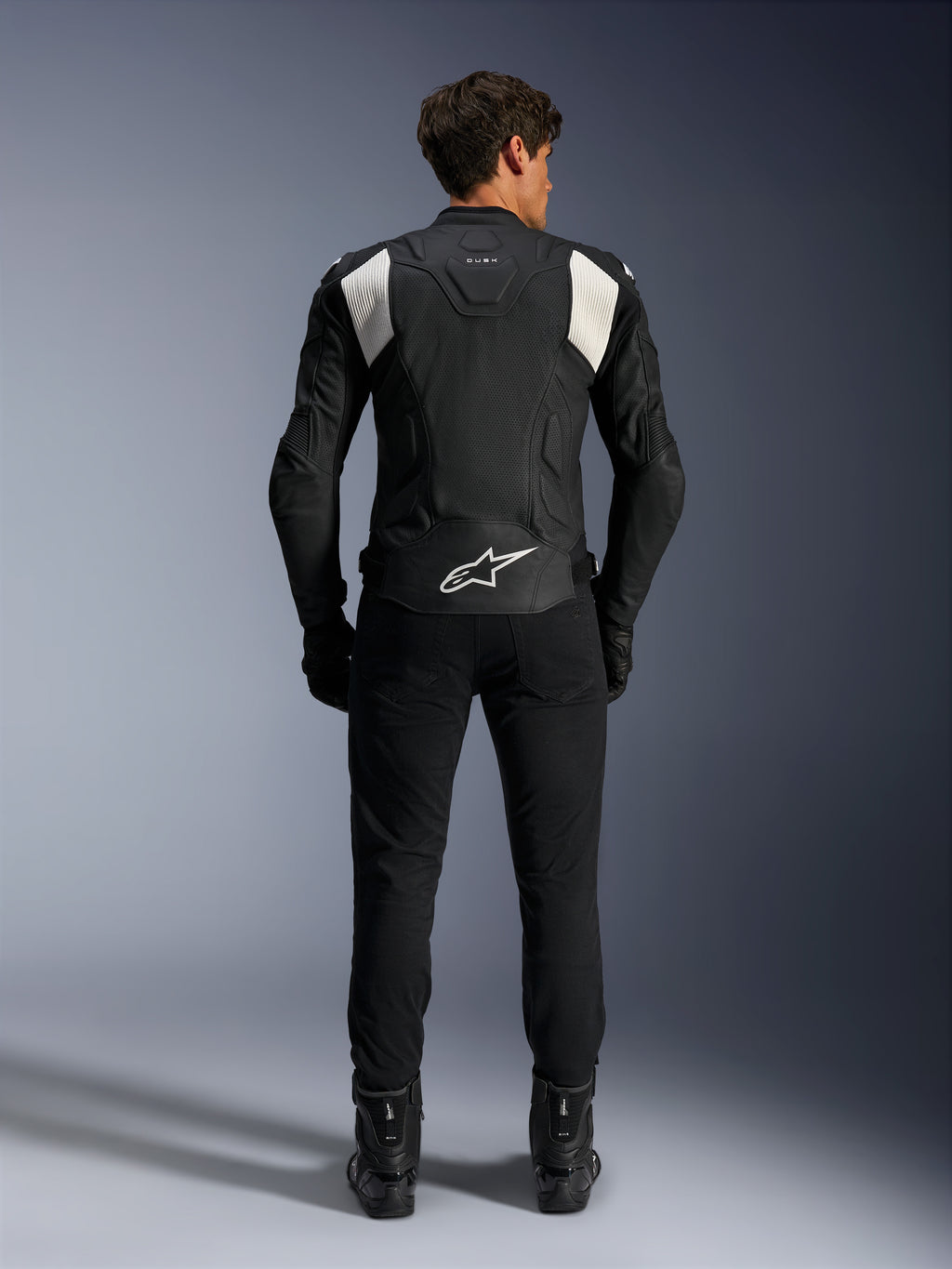 Chaqueta de cuero Alpinestars Dusk Airflow, en negro y blanco, vista trasera de un piloto con pantalones y botas negros, con cuero perforado para ventilación, paneles elásticos de acordeón blancos en los hombros y marca blanca en la parte baja de la espalda