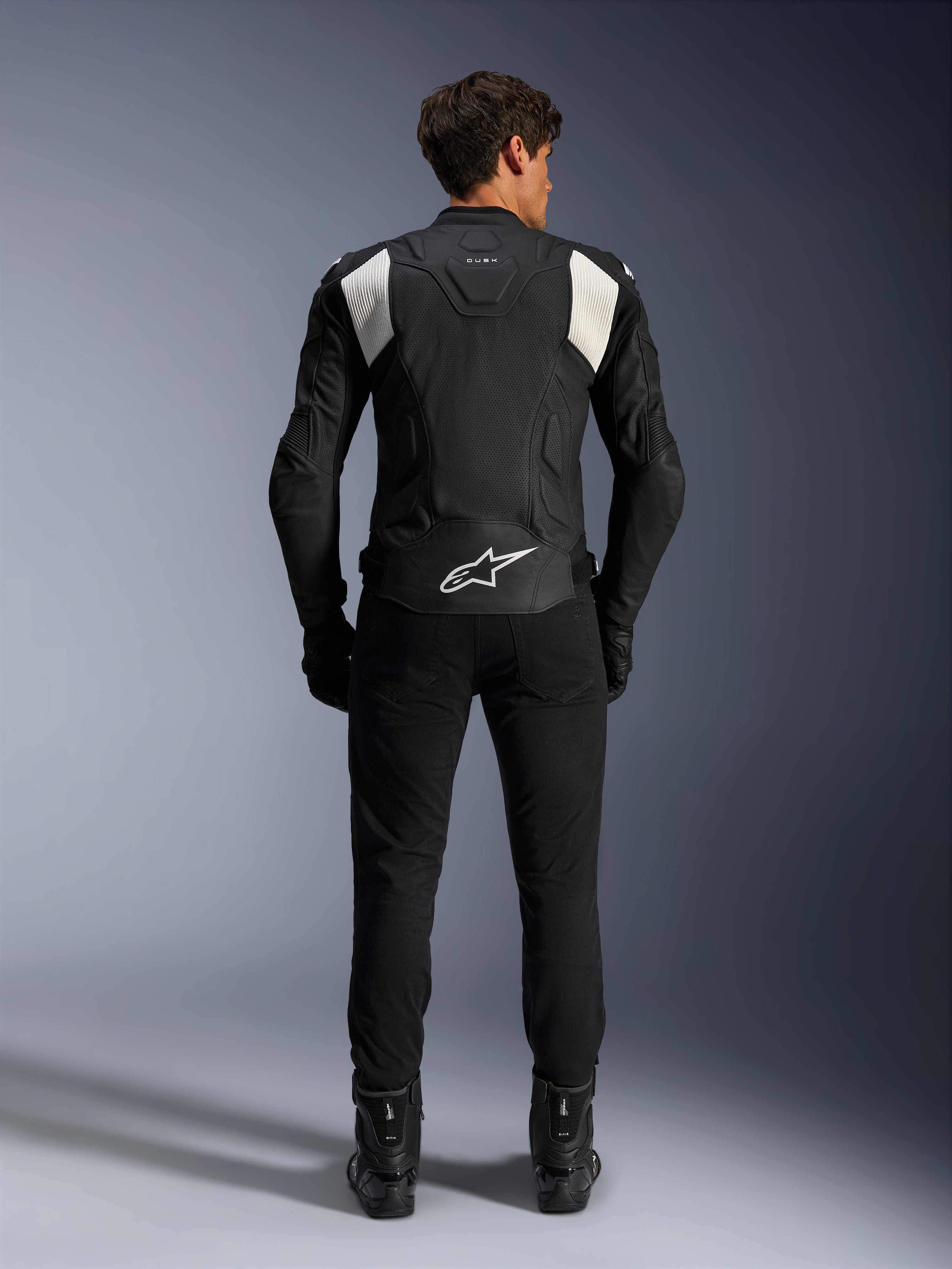 Chaqueta de cuero Alpinestars Dusk Airflow, en negro y blanco, vista trasera de un piloto con pantalones y botas negros, con cuero perforado para ventilación, paneles elásticos de acordeón blancos en los hombros y marca blanca en la parte baja de la espalda