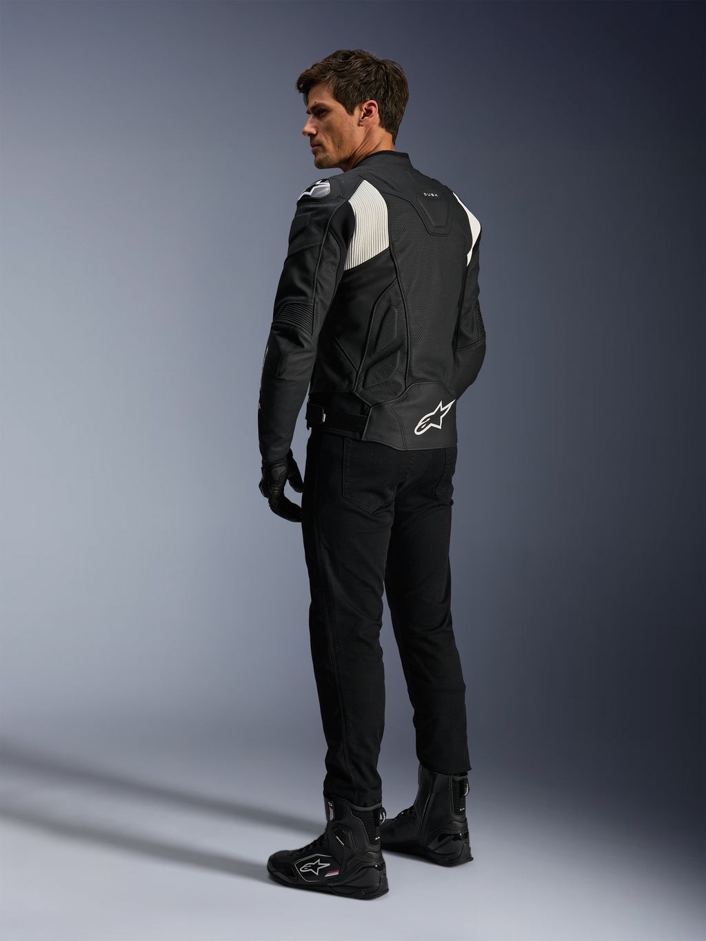 Chaqueta de cuero Alpinestars Dusk Airflow, negro y blanco, vista trasera de un modelo con la chaqueta de cuero perforado, pantalones y botas negras a juego, paneles elásticos de acordeón blancos y un logotipo llamativo en la parte baja de la espalda, equipamiento de conducción de moto deportiva