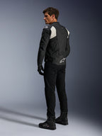 Chaqueta de cuero Alpinestars Dusk Airflow, negro y blanco, vista trasera de un modelo con la chaqueta de cuero perforado, pantalones y botas negras a juego, paneles elásticos de acordeón blancos y un logotipo llamativo en la parte baja de la espalda, equipamiento de conducción de moto deportiva