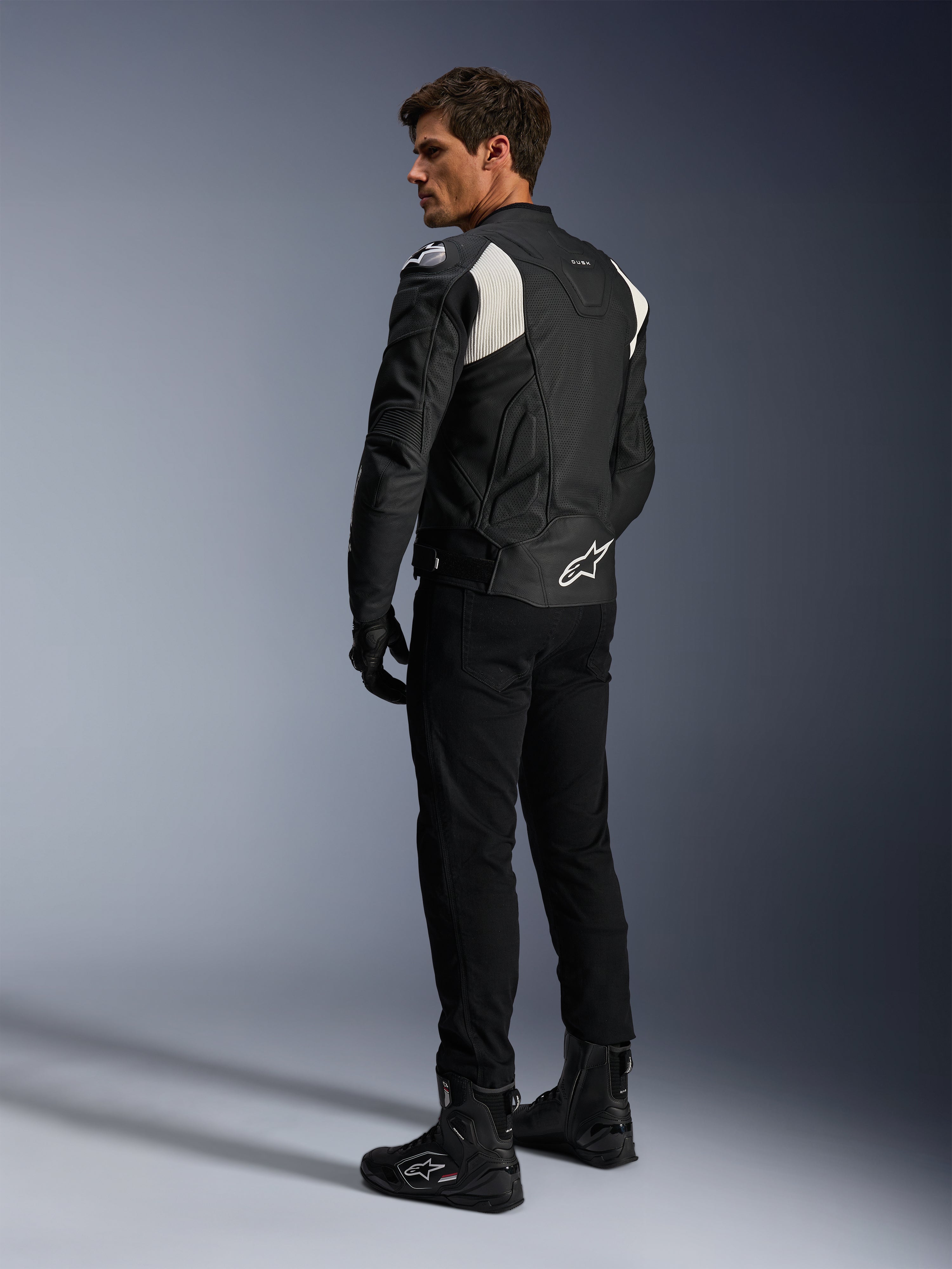 Chaqueta de cuero Alpinestars Dusk Airflow, negro y blanco, vista trasera de un modelo con la chaqueta de cuero perforado, pantalones y botas negras a juego, paneles elásticos de acordeón blancos y un logotipo llamativo en la parte baja de la espalda, equipamiento de conducción de moto deportiva
