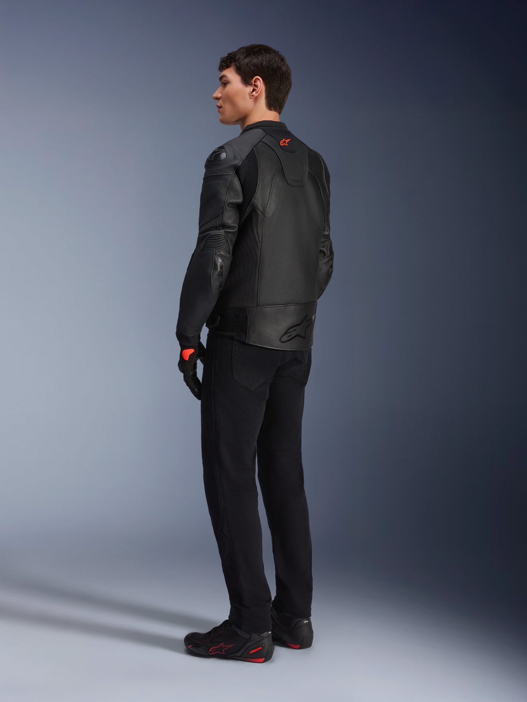 Chaqueta de cuero Alpinestars GP Plus R V4 Airflow, chaqueta deportiva de moto para hombre, totalmente negra, vista trasera de tres cuartos en un modelo, con paneles de cuero perforados, logotipo rojo en la espalda y protección de hombros integrada, combinada con pantalones negros y calzado de conducción